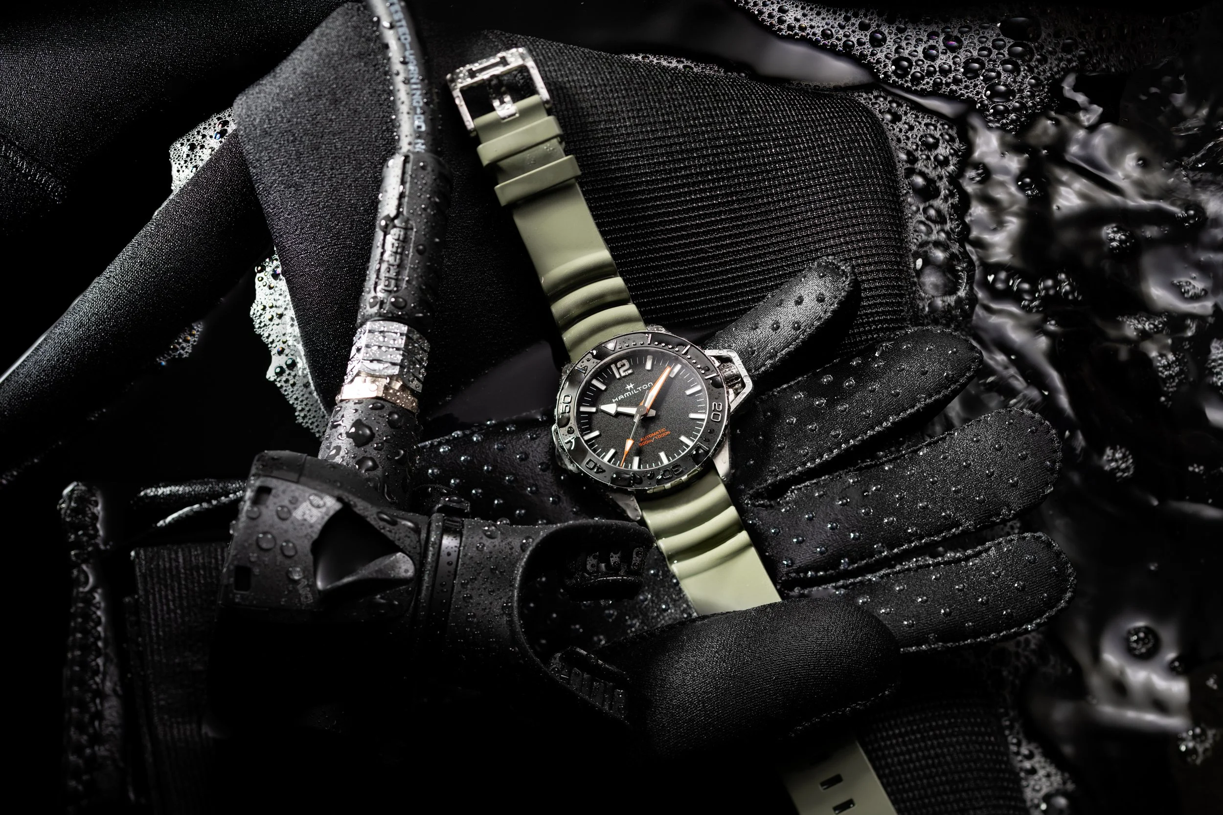 Photo publicitaire pour Hamilton: Une montre de plongée noire avec un bracelet vert, une combinaison de plongée noire avec de l'eau et des bulles, et des accessoires de plongée noirs, tous humides.