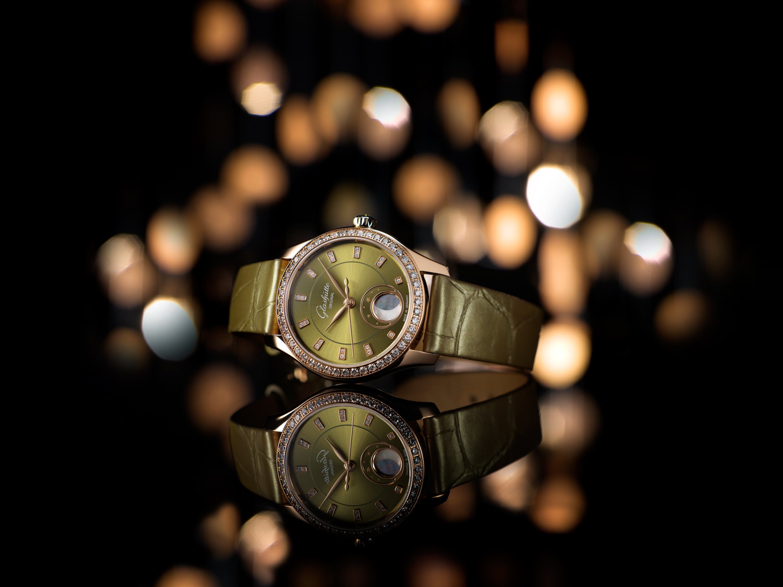 Photo de pub pour lancement montre Glashütte : Montre de luxe avec un cadran vert, un bracelet en cuir vert et un cadre entouré de petites pierres précieuses, reflétée sur une surface noire avec un fond flou de lumières dorées.