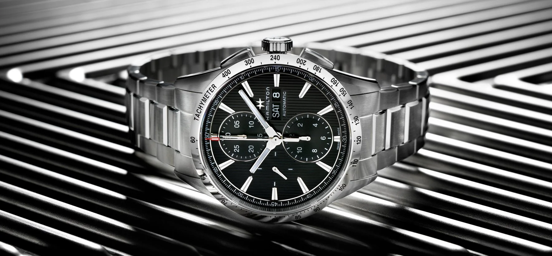Photo publicitaire pour Hamilton: Montre-bracelet en acier inoxydable avec cadran noir, chronographe, et affichage de la date, posée sur une grille métallique.