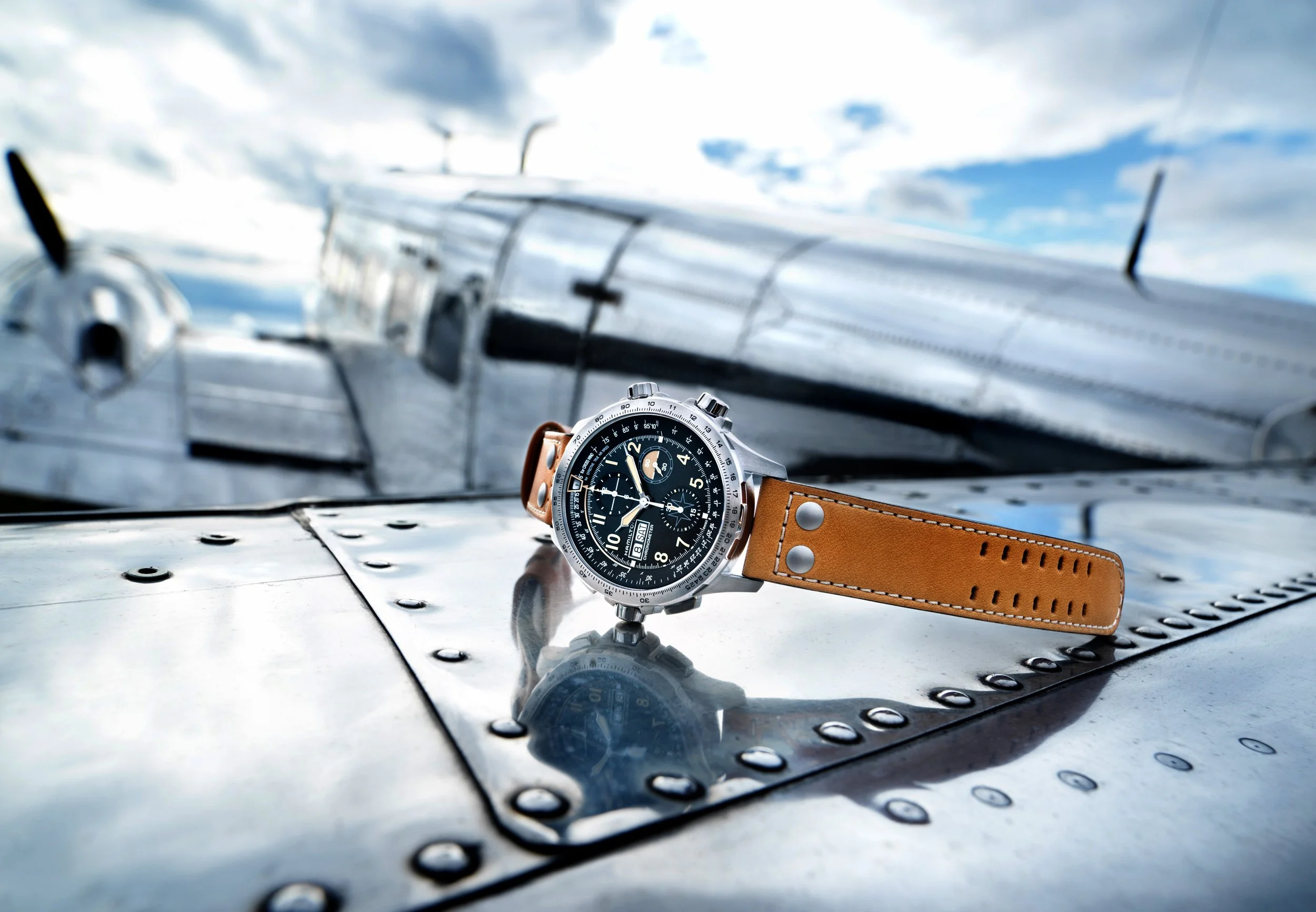Photo publicitaire pour Hamilton: Une montre-bracelet avec un bracelet en cuir marron posée sur une surface métallique, avec un fond flou d'un avion en métal contre un ciel partiellement nuageux.