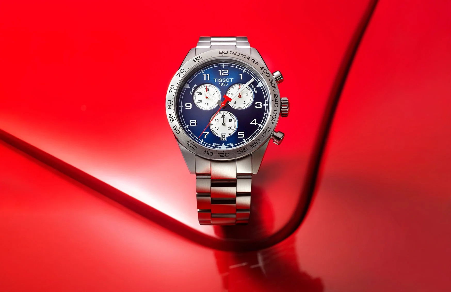 Visuel pub: Montre Tissot chronographe avec un cadran bleu, bracelet en acier, posé sur capot de voiture rouge.
