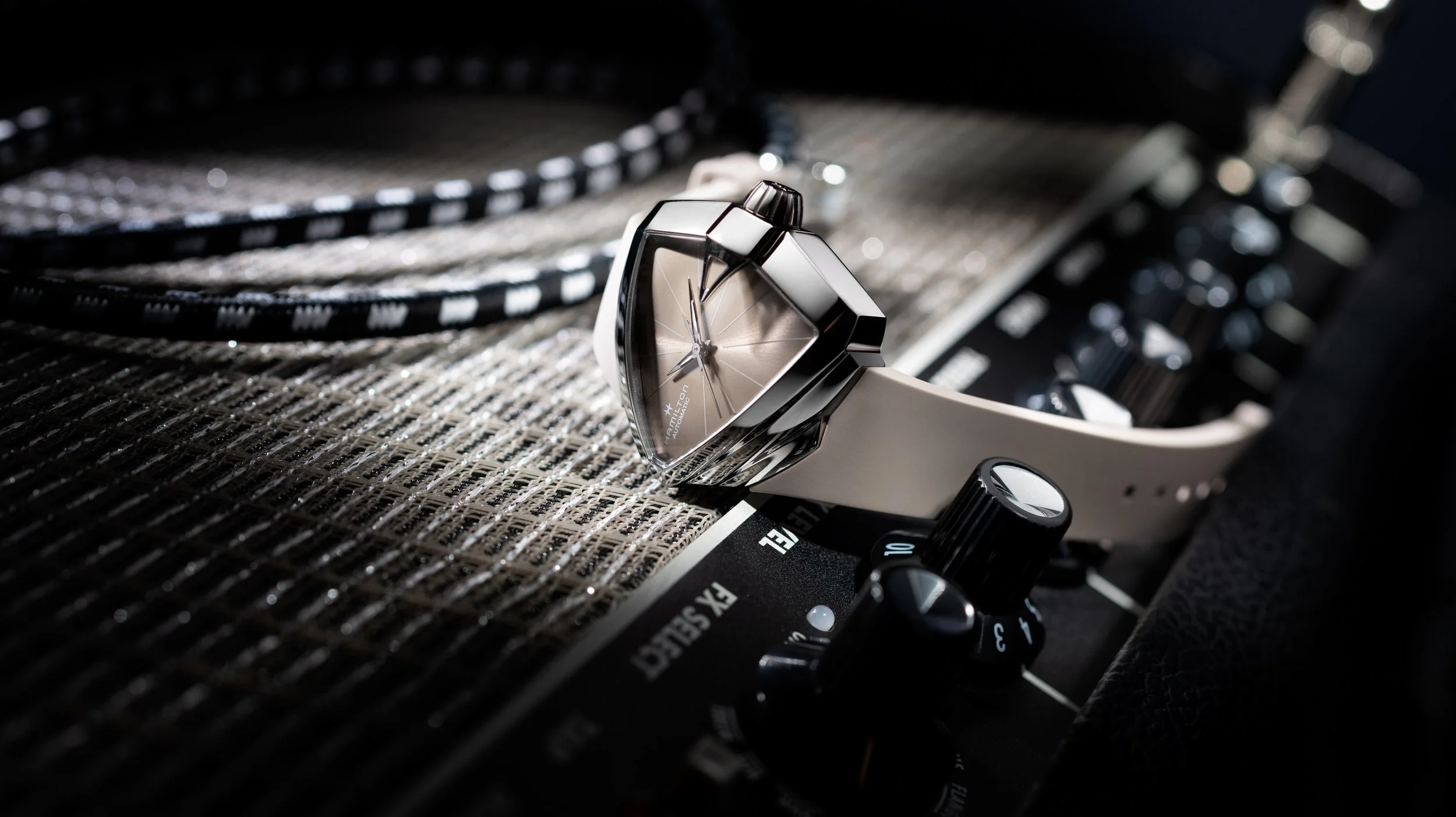 Photo publicitaire pour Hamilton: Une montre posée sur un amplificateur audio avec des commandes de réglage visibles, l'image est sombre avec un éclairage ciblé sur la montre et l'amplificateur.