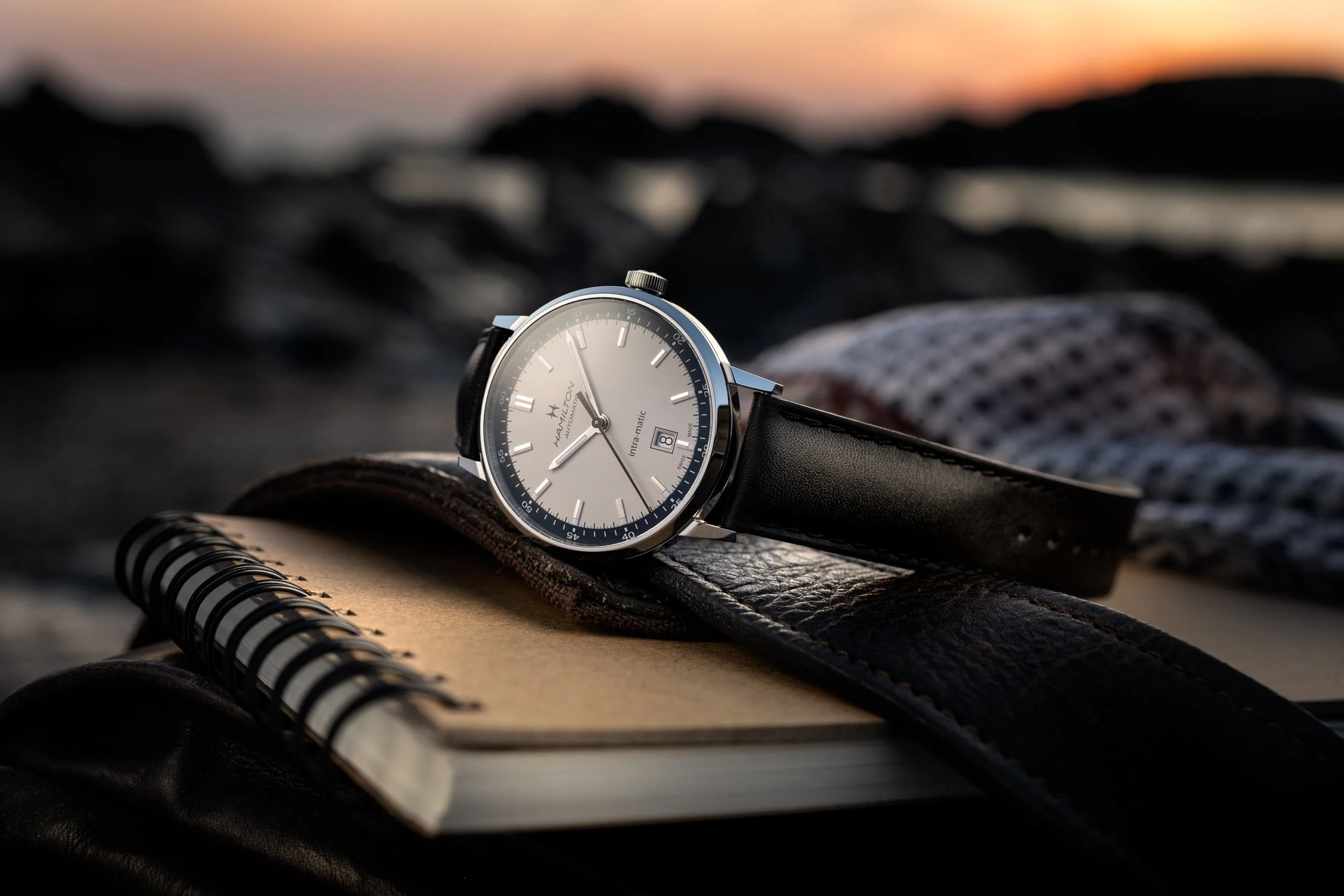 Photo publicitaire pour Hamilton: Montre élégante avec cadran argenté, posée sur un carnet, en plein air au coucher du soleil.