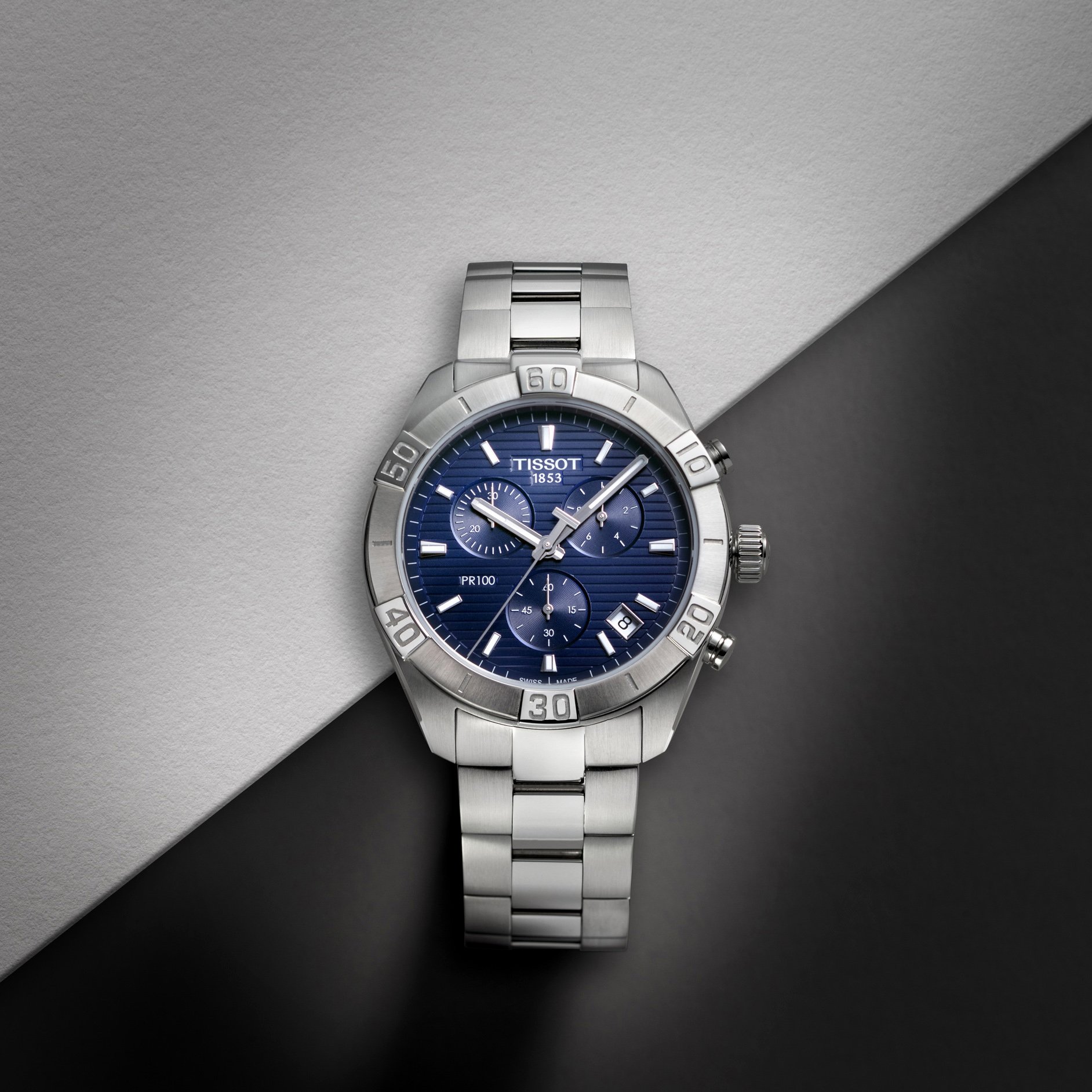 Visuel pub pour Tissot: Montre en acier inoxydable avec un cadran bleu, posée sur un fond bicolore gris et noir.