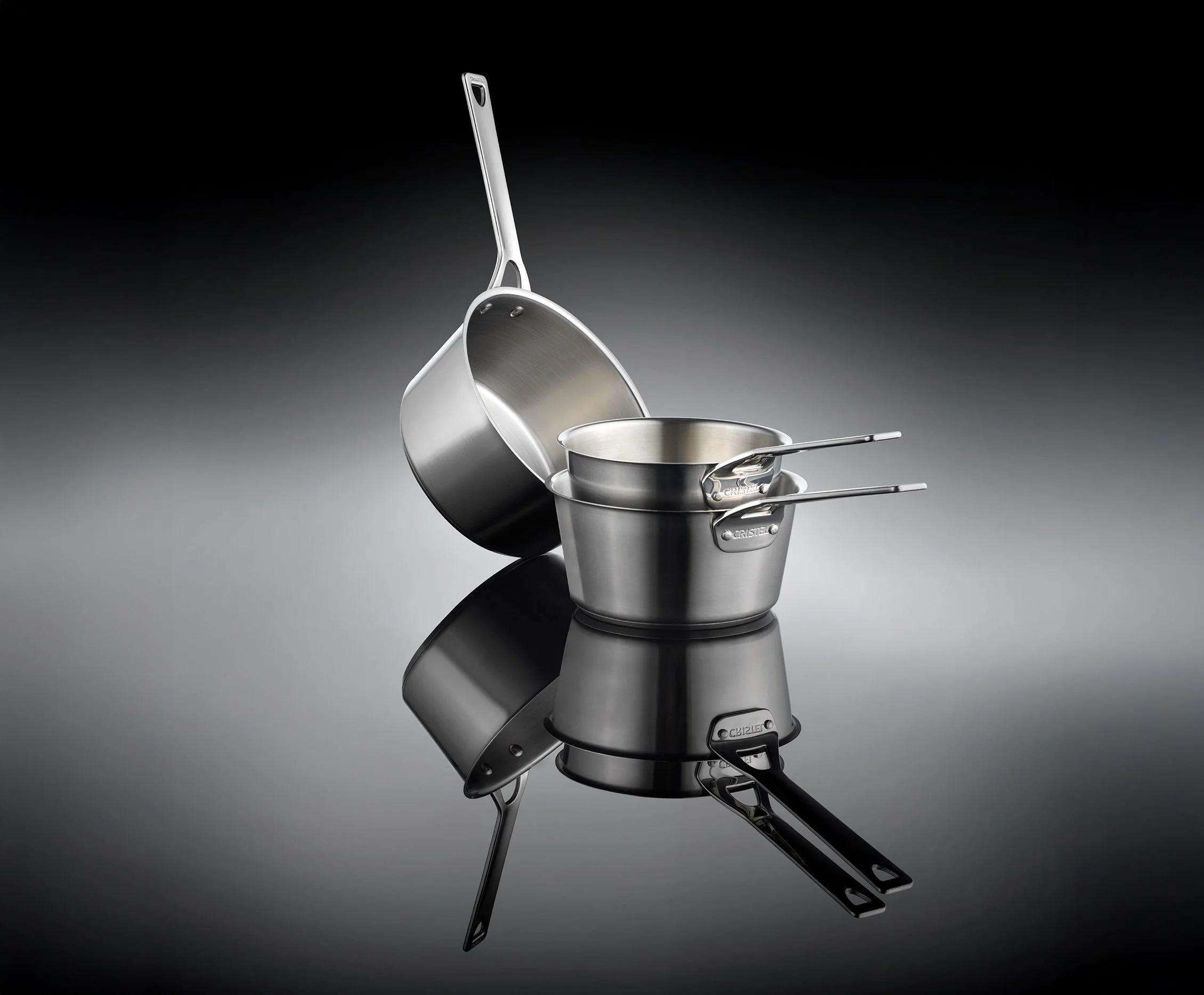 Set de casseroles de la marque Cristel en acier inoxydable, comprenant deux casseroles empilées avec poignées, et une poêle suspendue, avec un reflet sur une surface noire et un fond sombre allumé par un éclairage dirigé.