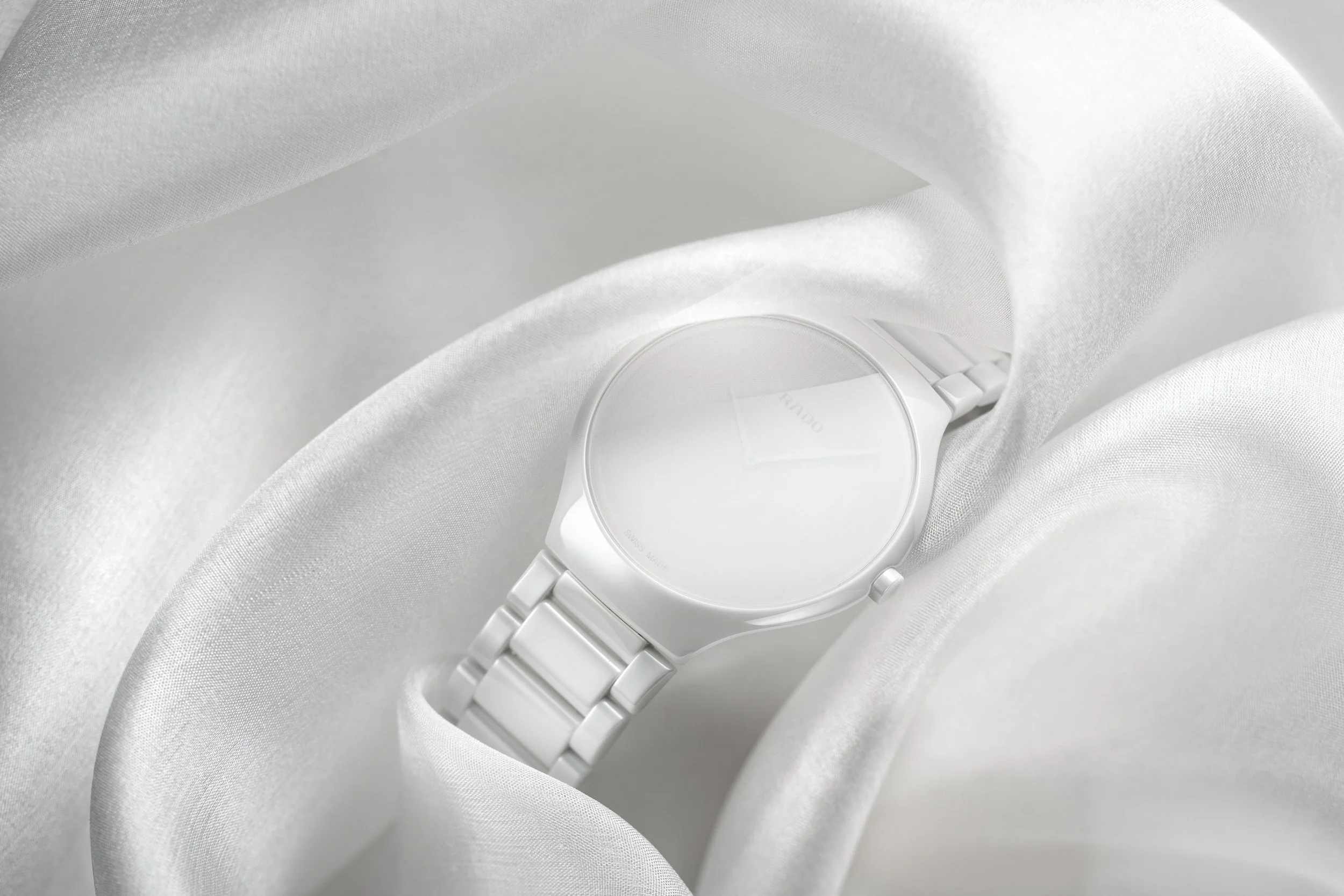 Visuel pub pour Rado : Montre blanche posée sur des tissus satinés blancs.