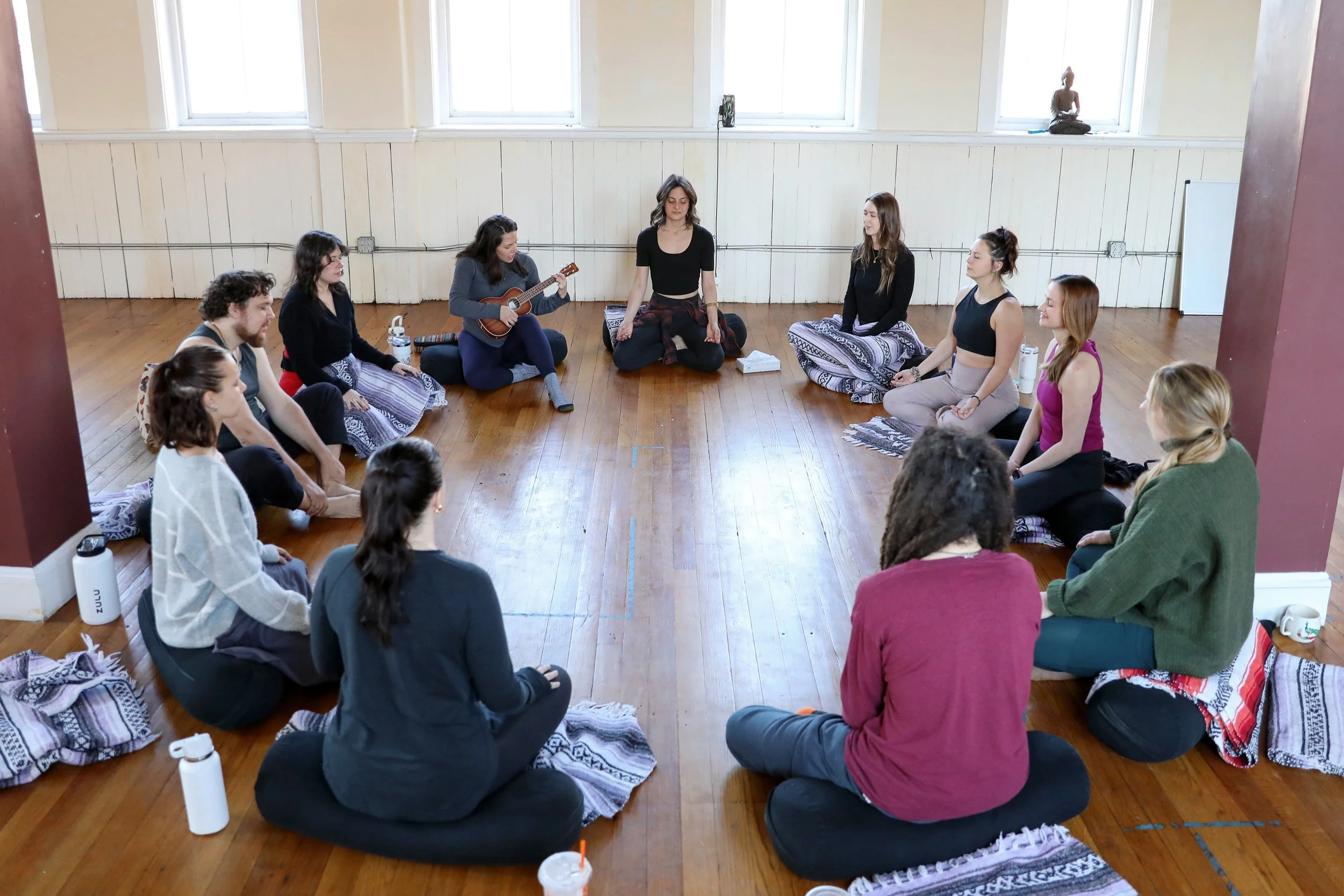 A group of women participating in a yoga or stretching class in a bright and sunny yoga studio. One woman is extending her arms forward while others are sitting or lying on the mats, learning to how make hands-on adjustments.