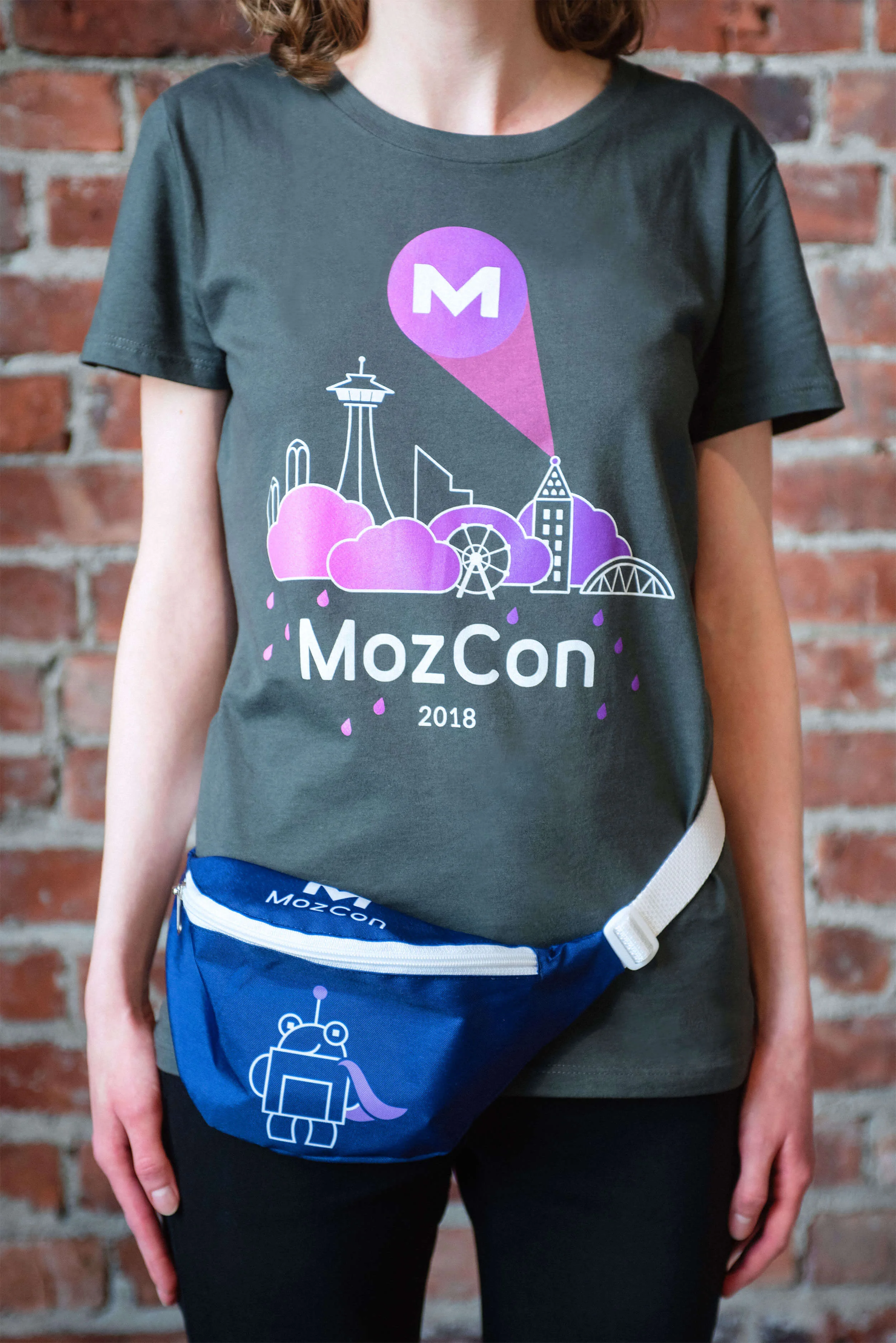 MozCon 2018 swag: Mozopolis cityscape graphic tee featuring the Roger signal (AKA the MozCon logo) projected from the illustrated Smith Tower, and purple Super Roger fanny pack.