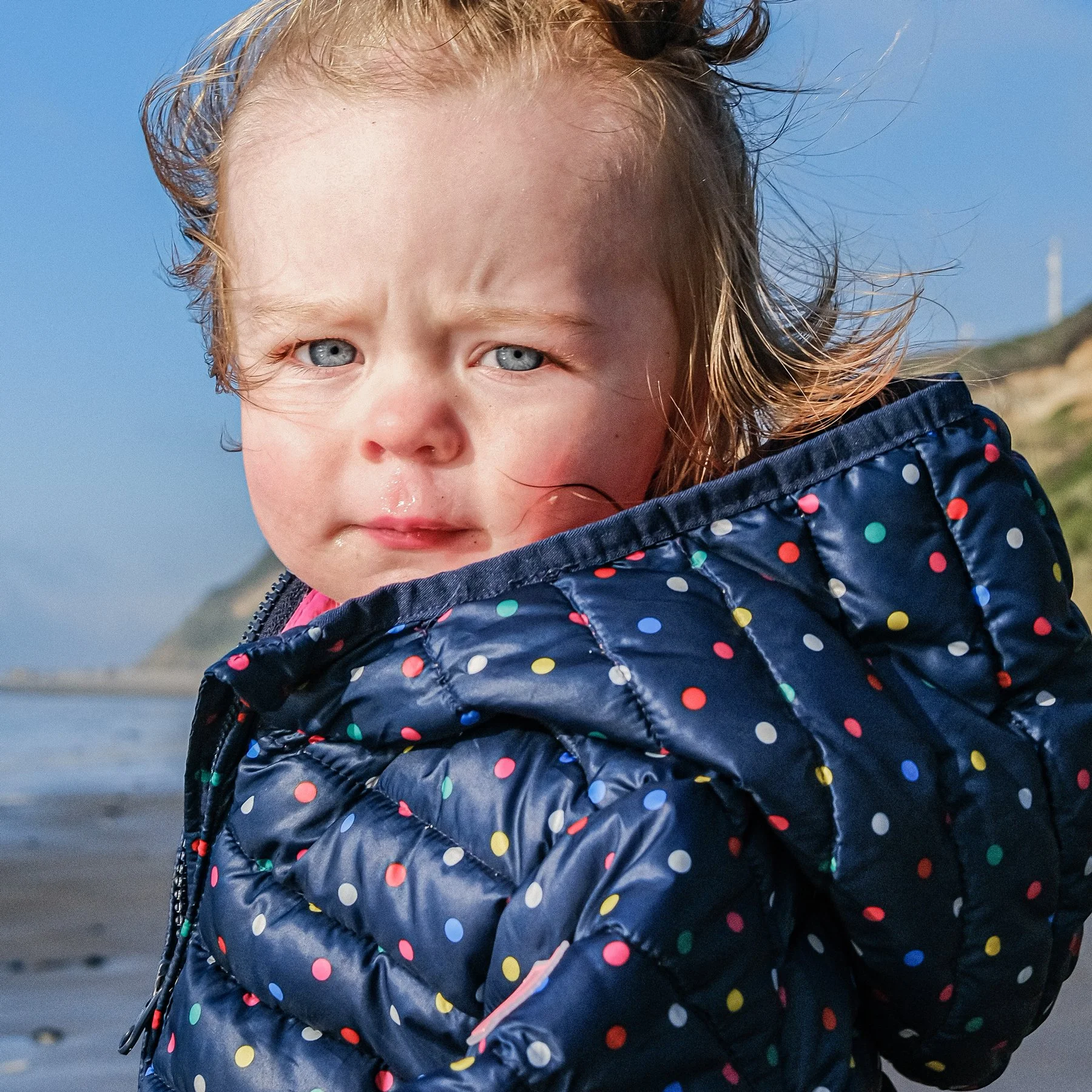 A young girl with wet hair and a sad expression, wearing a blue polka dot jacket, standing on a beach with cliffs and a cloudy sky in the background.