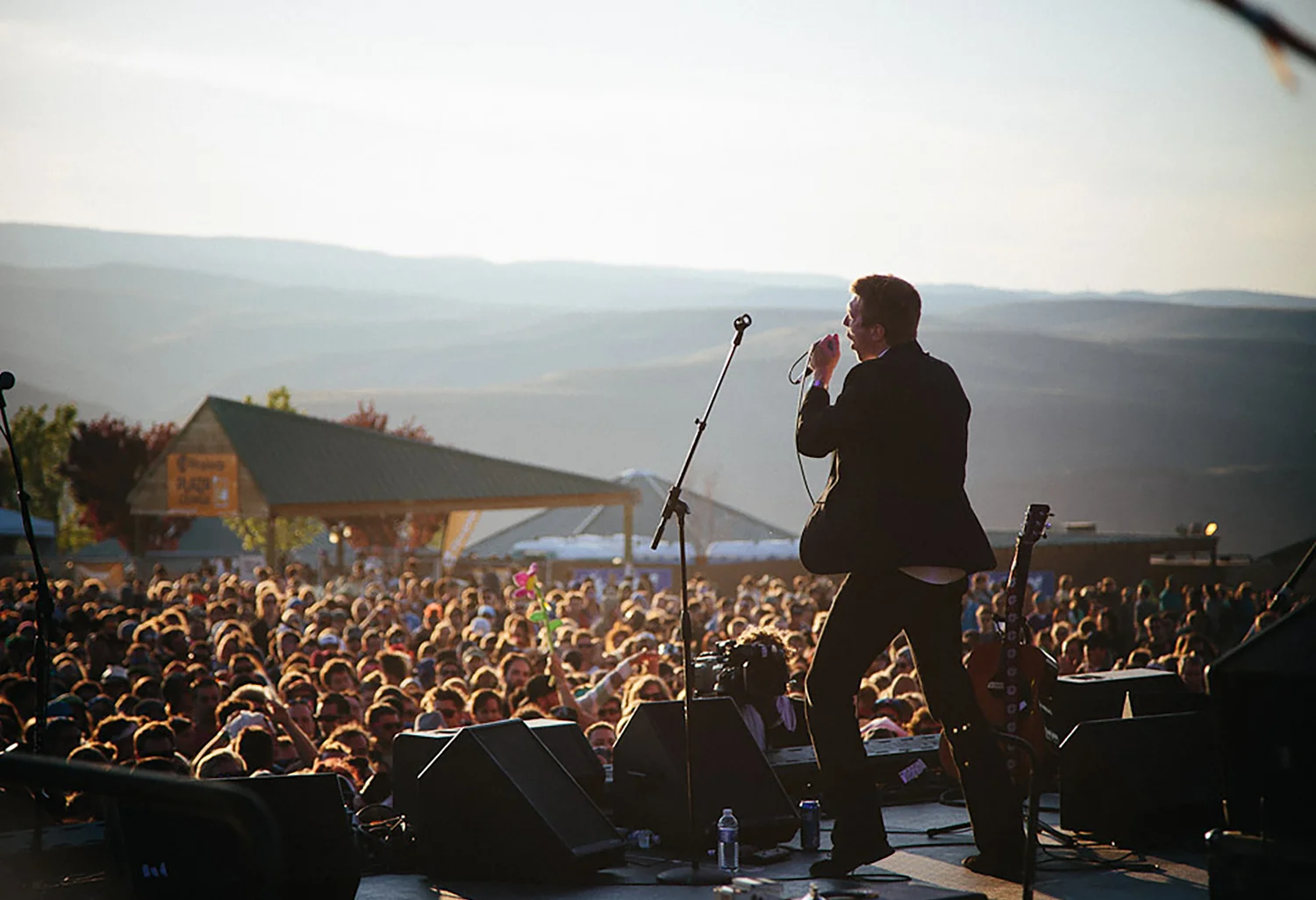 Male singer on stage performing at an outdoor concert with a large crowd in the background and mountains in the distance.