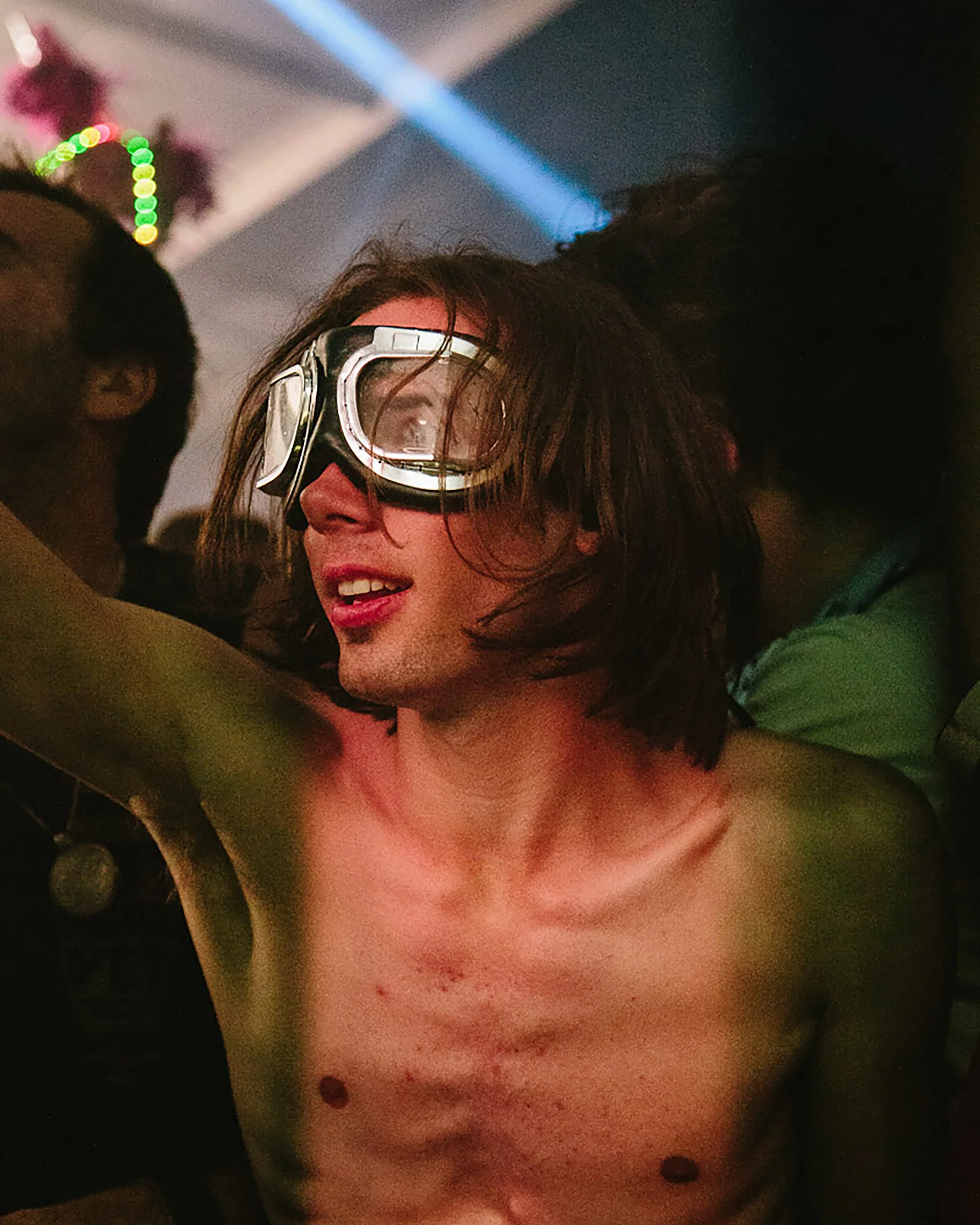 A person with shoulder-length brown hair and wearing swimming goggles is smiling at a party or event with colorful lights in the background. They are shirtless and appear to have some scratches or marks on their chest.