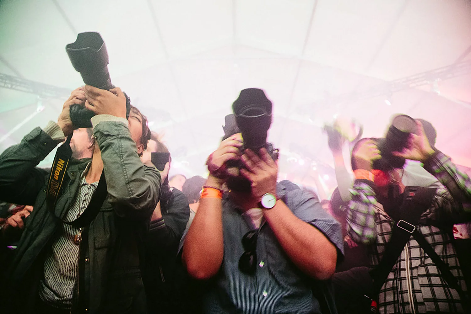 Group of people at a concert, holding cameras and phones, with some wearing black masks, in an indoor venue with colorful lighting.