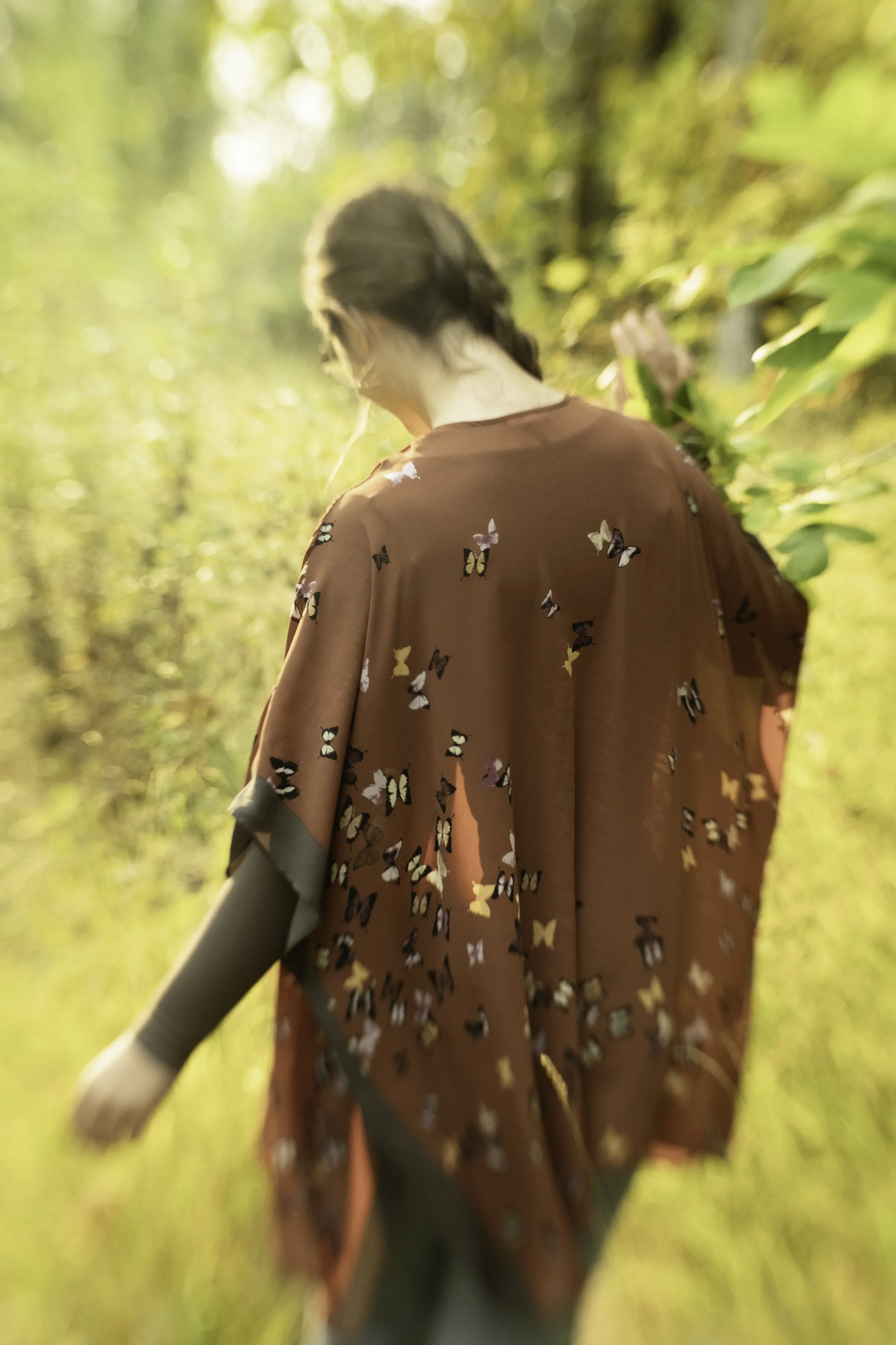 A woman walking through a sunlit outdoor forest or park area, wearing a brown shawl or garment with butterfly patterns.