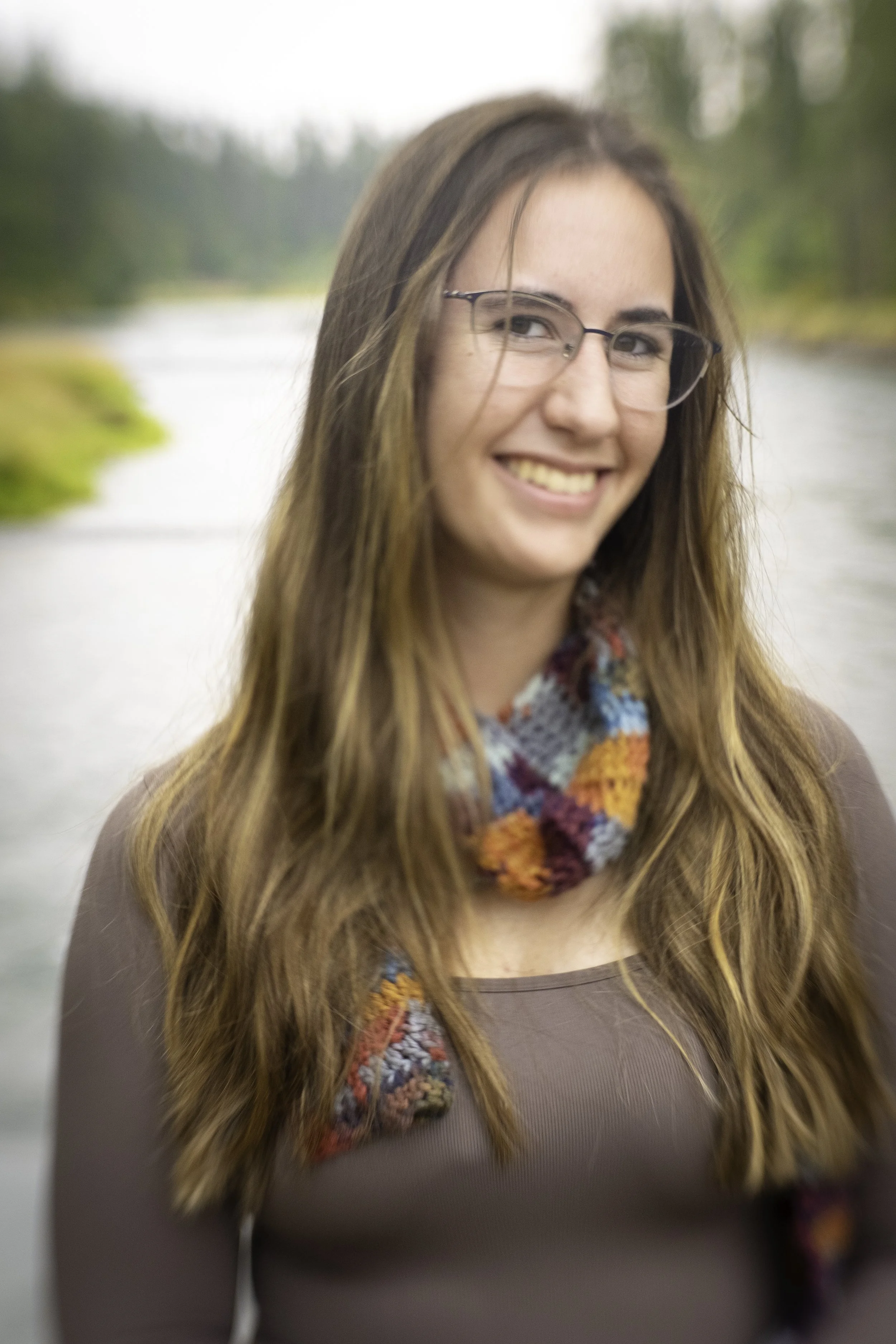 A woman with long, wavy brown hair, glasses, and a colorful scarf stands outdoors near a river, smiling at the camera.
