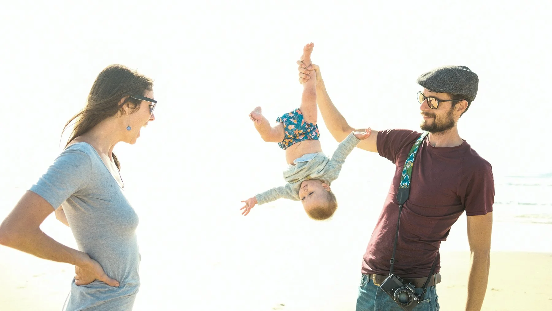 A young girl hanging upside down, held by a man, while a woman stands nearby on a beach with the ocean in the background.