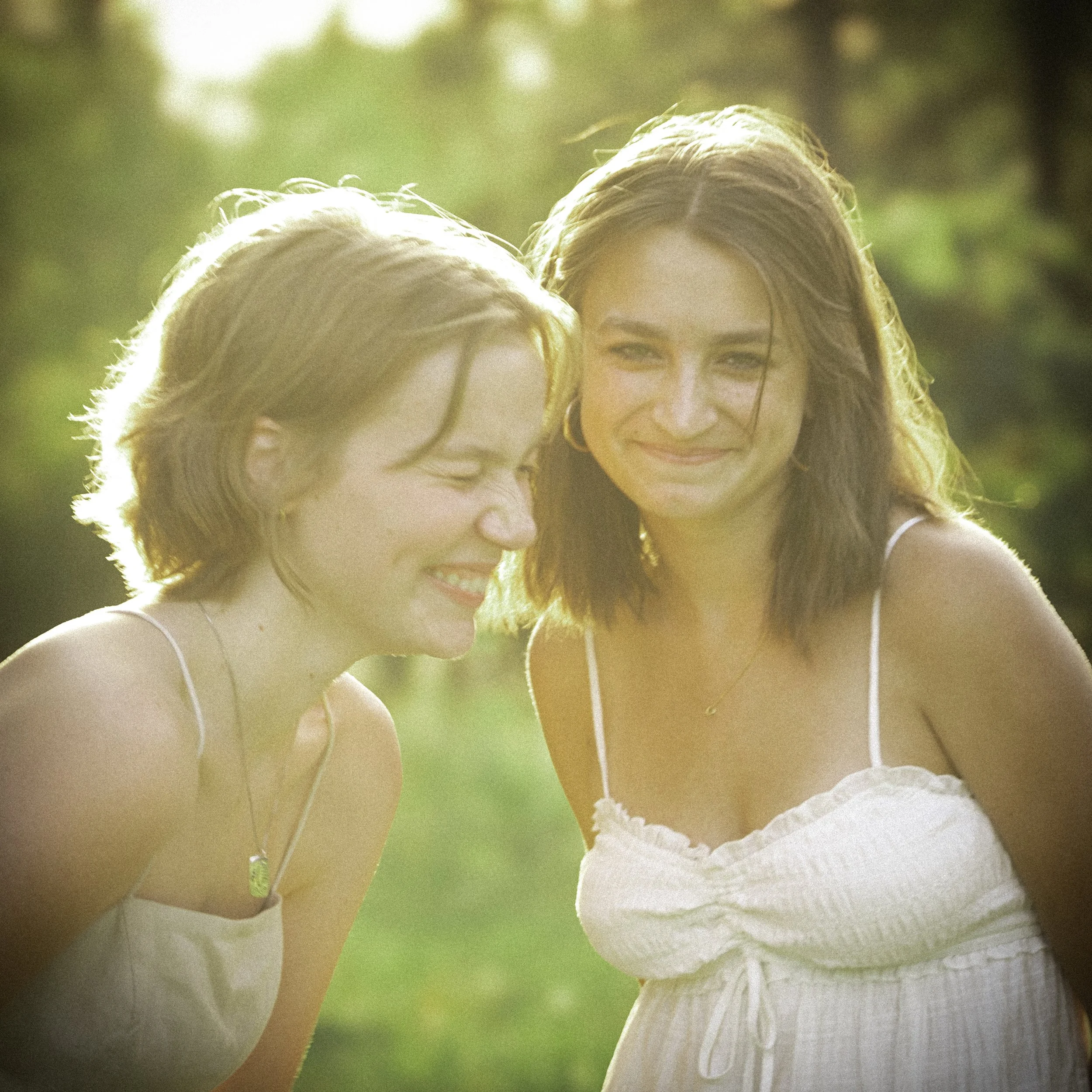 Two young women smiling and laughing outdoors in sunlight, wearing light-colored sleeveless dresses.