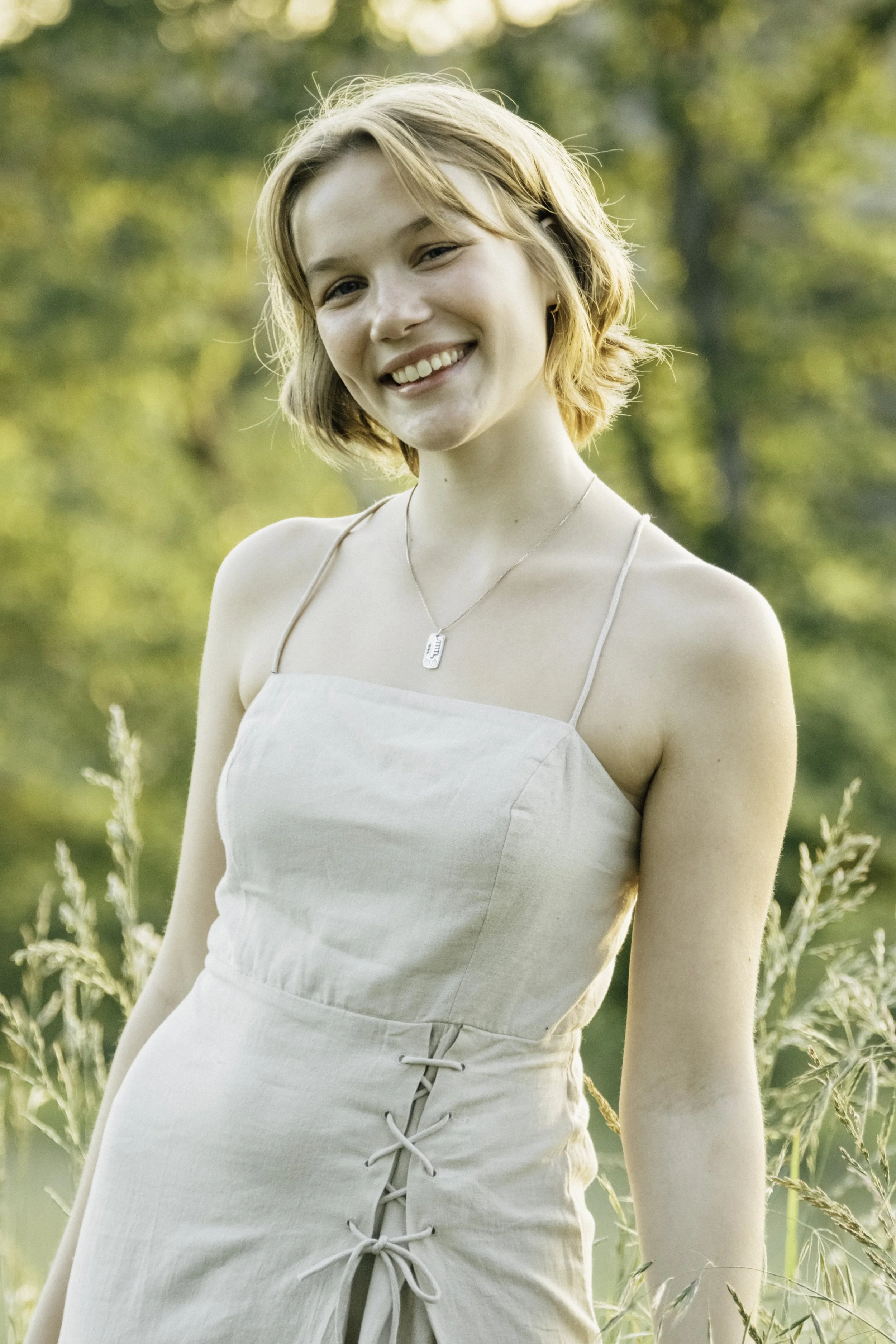 A young woman with short, wavy blonde hair smiling outdoors during daytime, wearing a light-colored sleeveless dress with a lace-up detail on the side and a necklace.