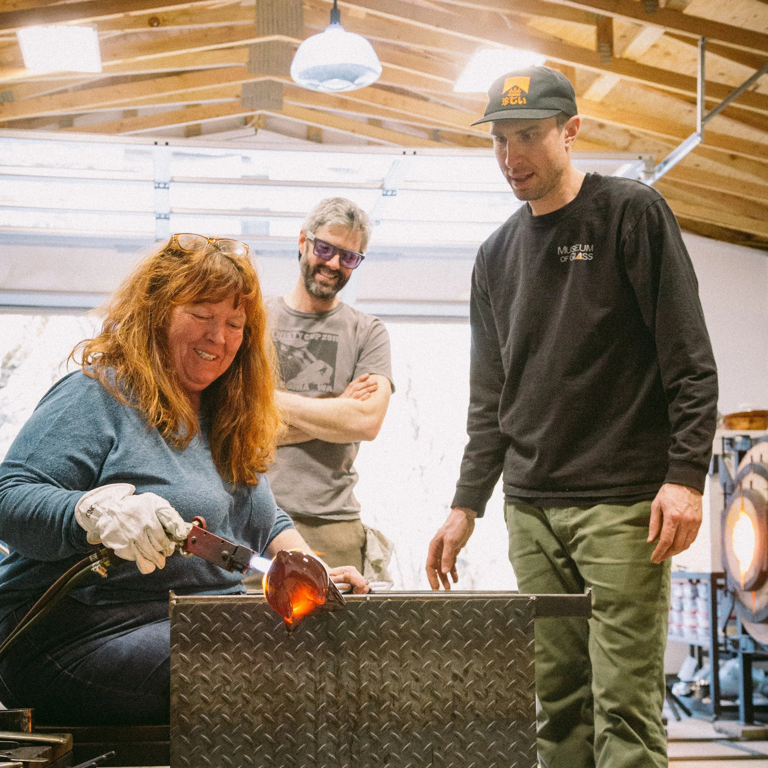 Three people in a glassblowing workshop observe as a woman shapes molten glass into a glass object. The woman, with red hair, is smiling and wearing gloves. A man with gray hair, beard, and purple glasses stands behind her with arms crossed, smiling.