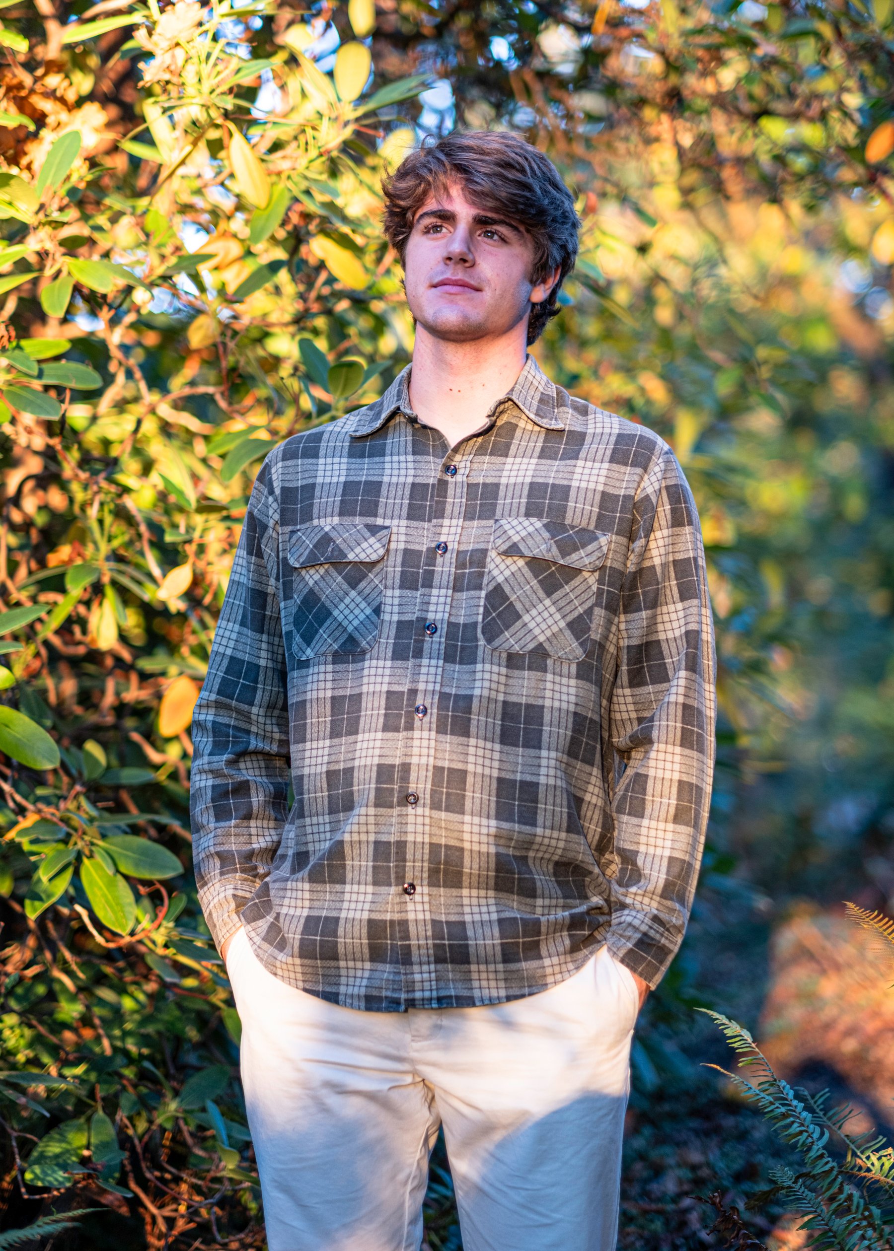 A young man with brown, wavy hair standing outdoors in front of green and yellow foliage, wearing a plaid shirt and white pants.