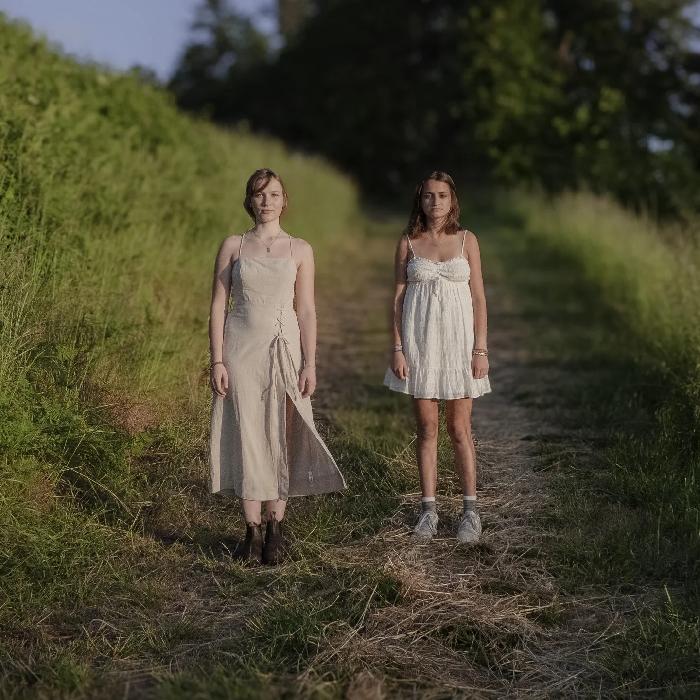 Two young women standing on a grassy dirt path in a rural area during sunset, surrounded by green trees and bushes, wearing casual summer dresses.