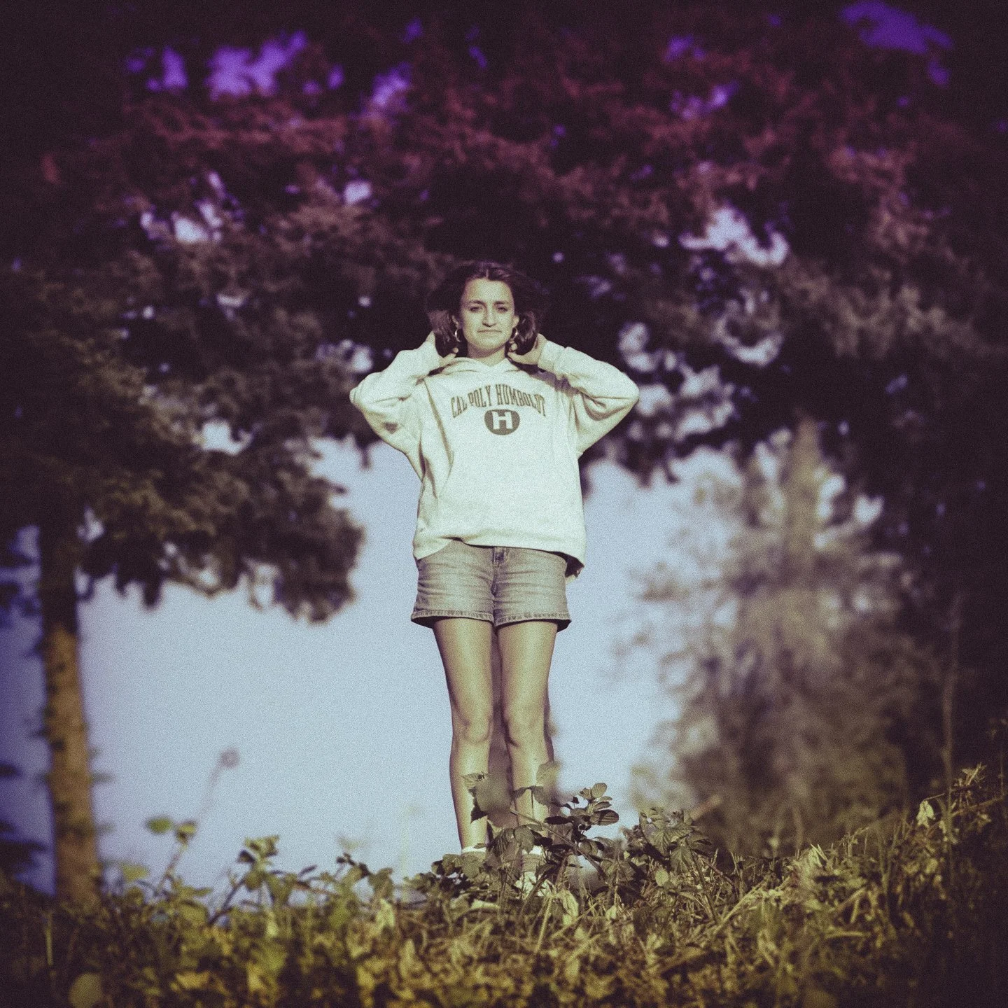 A young woman standing outdoors on grass, wearing a cream-colored hoodie and denim shorts, with trees and sky in the background.