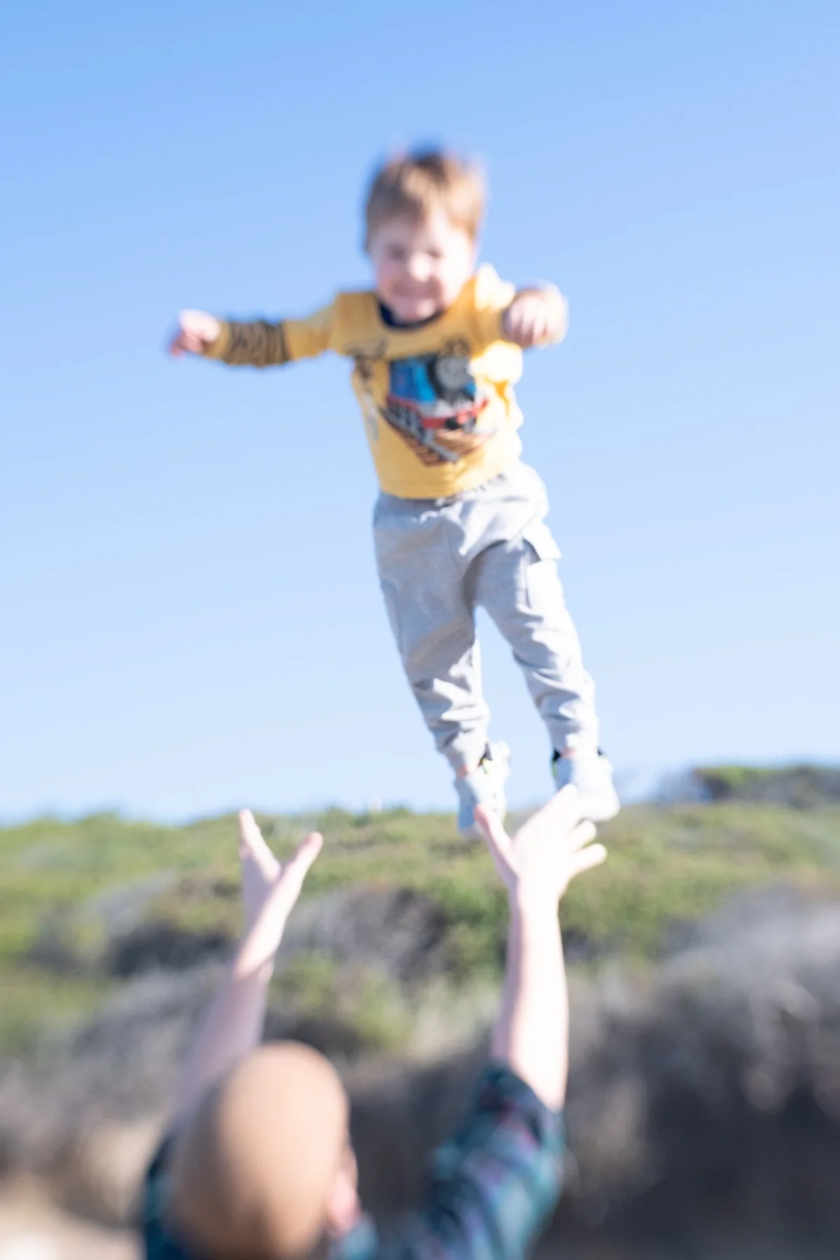 A young child being tossed into the air by an adult, outdoors on a sunny day, with clear blue sky and grassy terrain in the background.