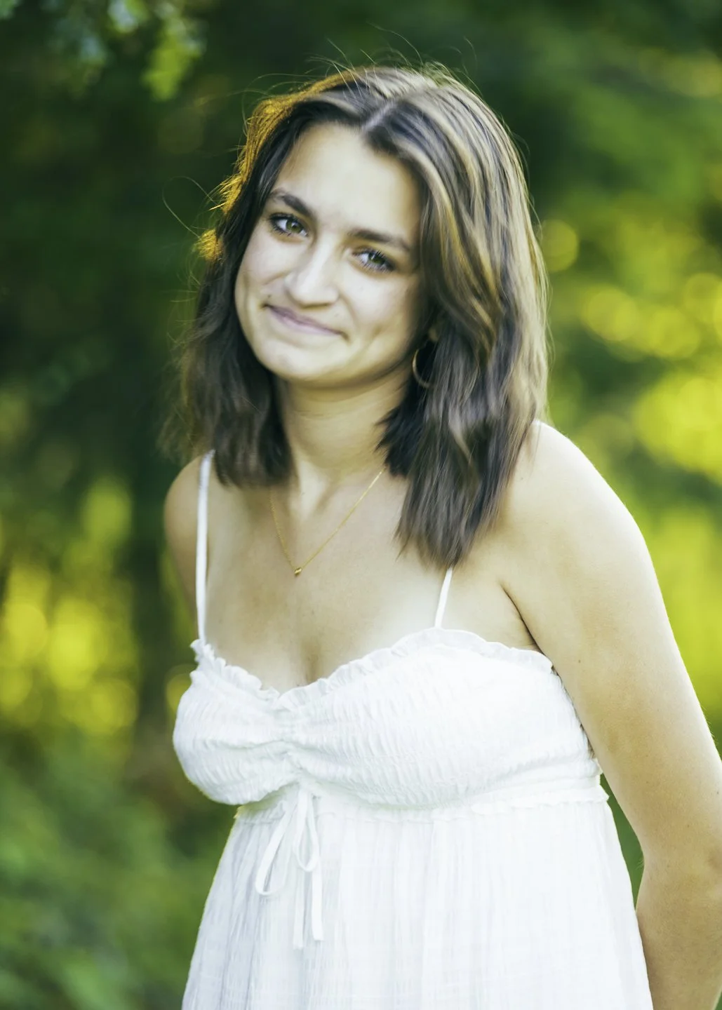 A woman with shoulder-length wavy brown hair wearing a white sleeveless dress, standing outdoors with blurred green foliage in the background, smiling softly.