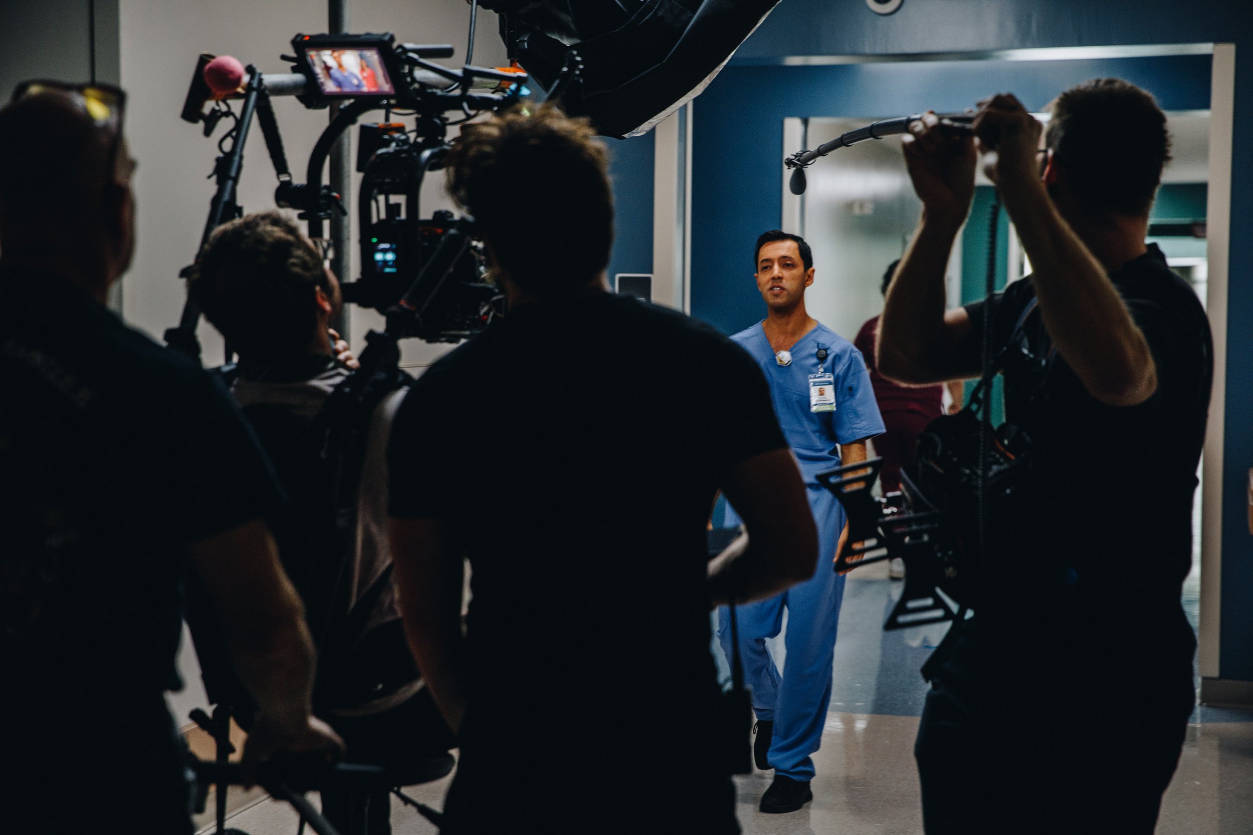 A group of film crew members filming a scene in a hospital corridor, featuring a man in blue medical scrubs speaking to the camera.