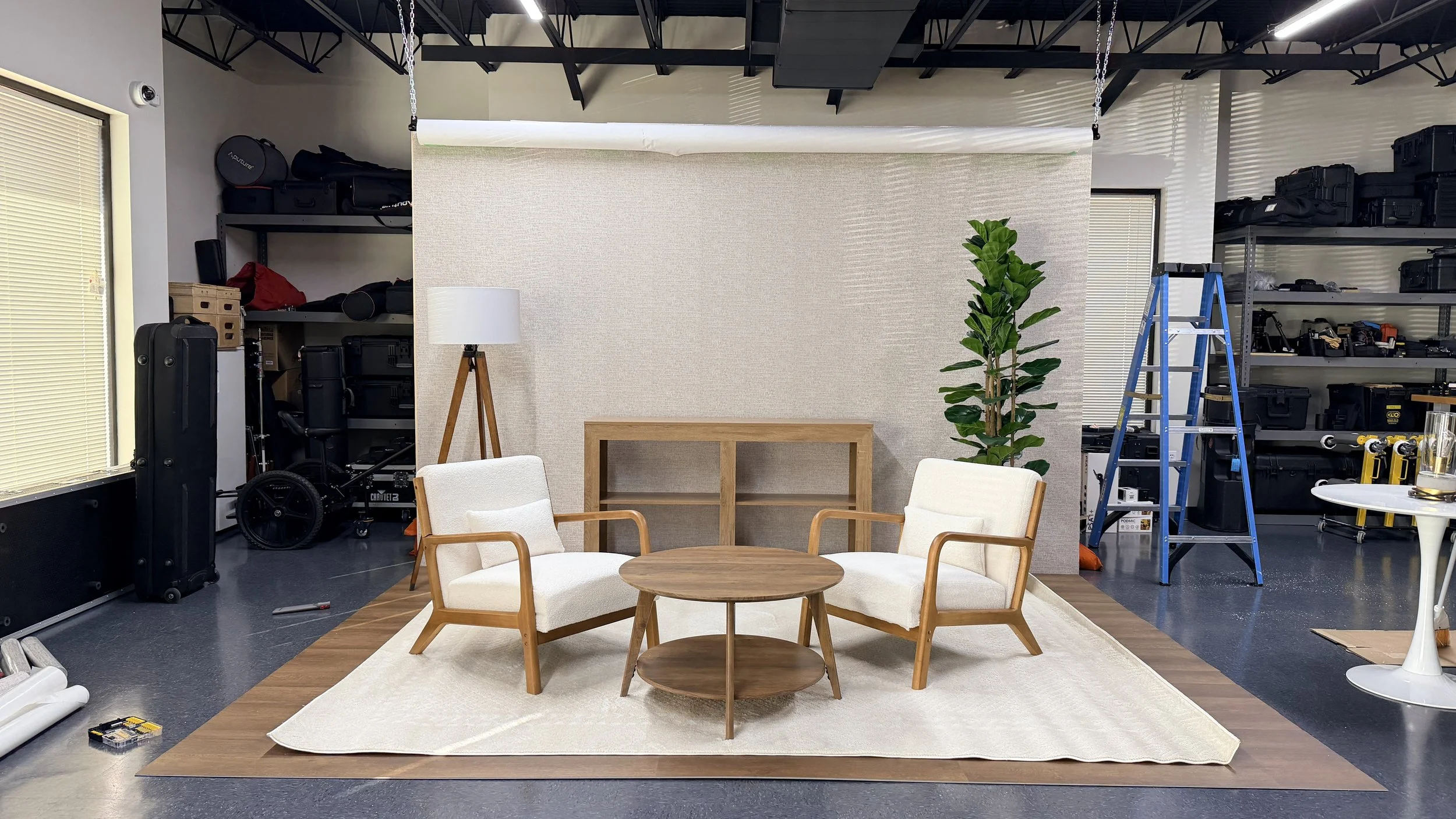 A cozy indoor scene setup with two white cushioned armchairs with wooden frames, a round wooden coffee table, and a standing floor lamp with a white shade. A tall green potted plant is on the right, and a wooden shelf is in the background. The area is surrounded by industrial shelving with equipment and supplies, with a ladder on the right side, and the setup is on a beige rug on a dark floor with natural light coming through windows with blinds.