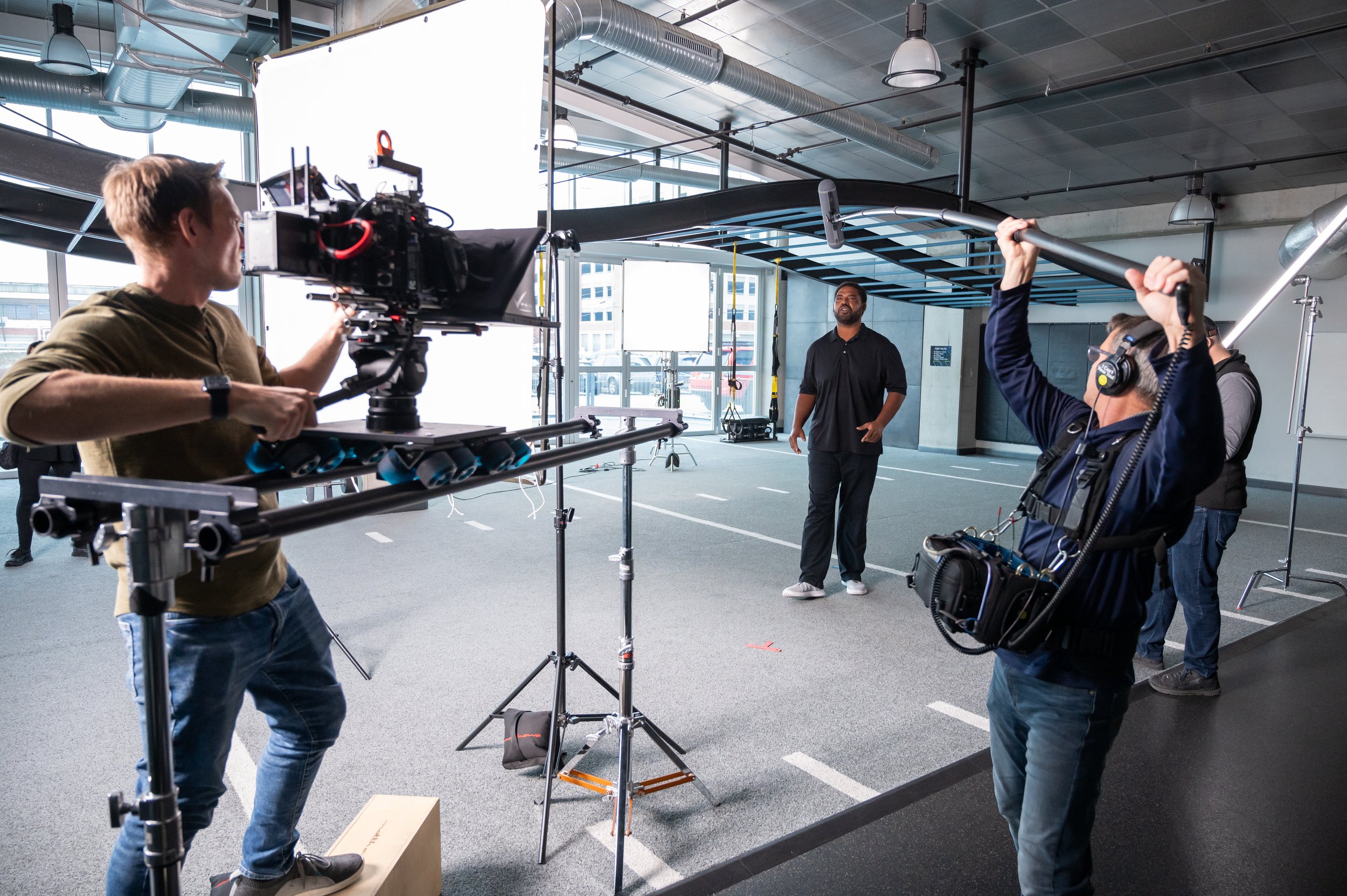 A film crew shoots a scene with an actor in an indoor setting resembling a parking garage. The crew uses professional camera equipment, boom mic, and headphones, with a director or interviewer watching the actor.