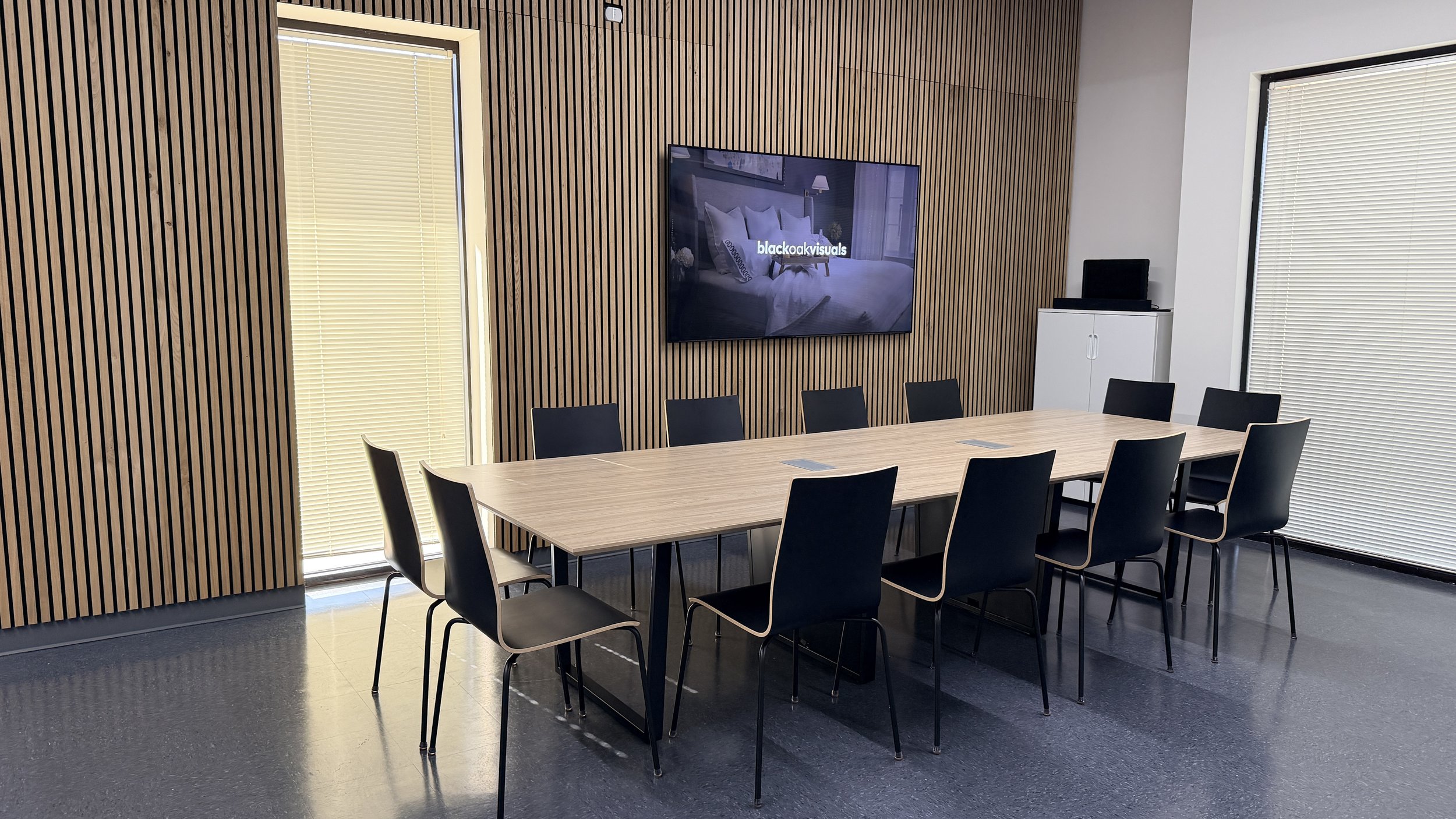 Empty conference room with a long wooden table, black chairs, wood-paneled wall, large window blinds, a mounted TV showing a bedroom, and a small white cabinet with a black object on top.