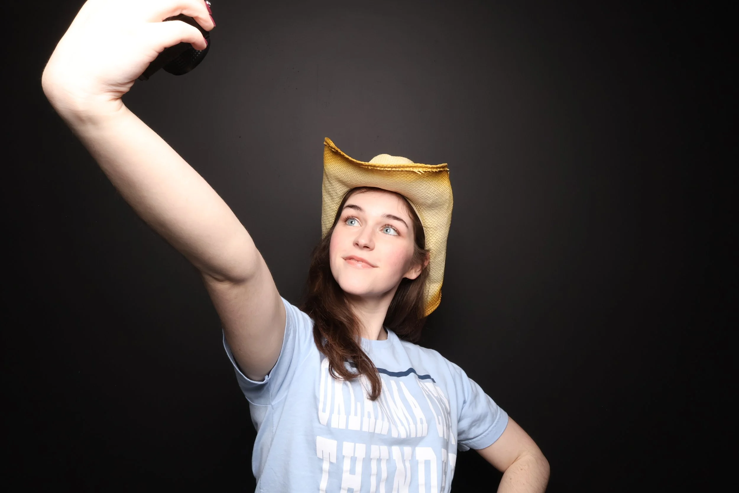 A young woman with brown hair and blue eyes wearing a light blue T-shirt takes a selfie with a smartphone while wearing a vintage-style cowboy hat against a dark background.