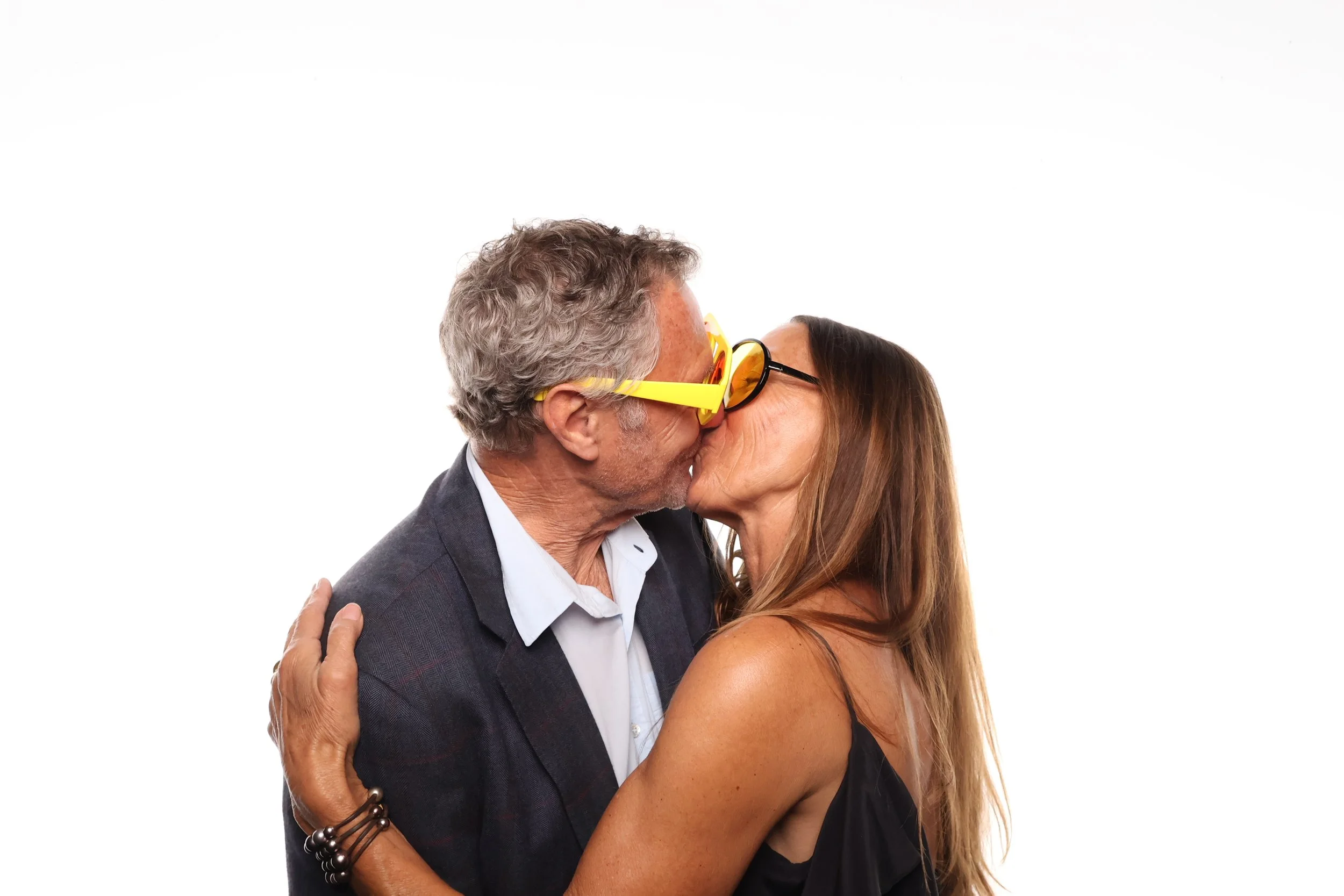 An older man wearing yellow sunglasses and a dark blazer is kissing a woman with long brown hair and black dress, with their arms around each other, against a plain white background.