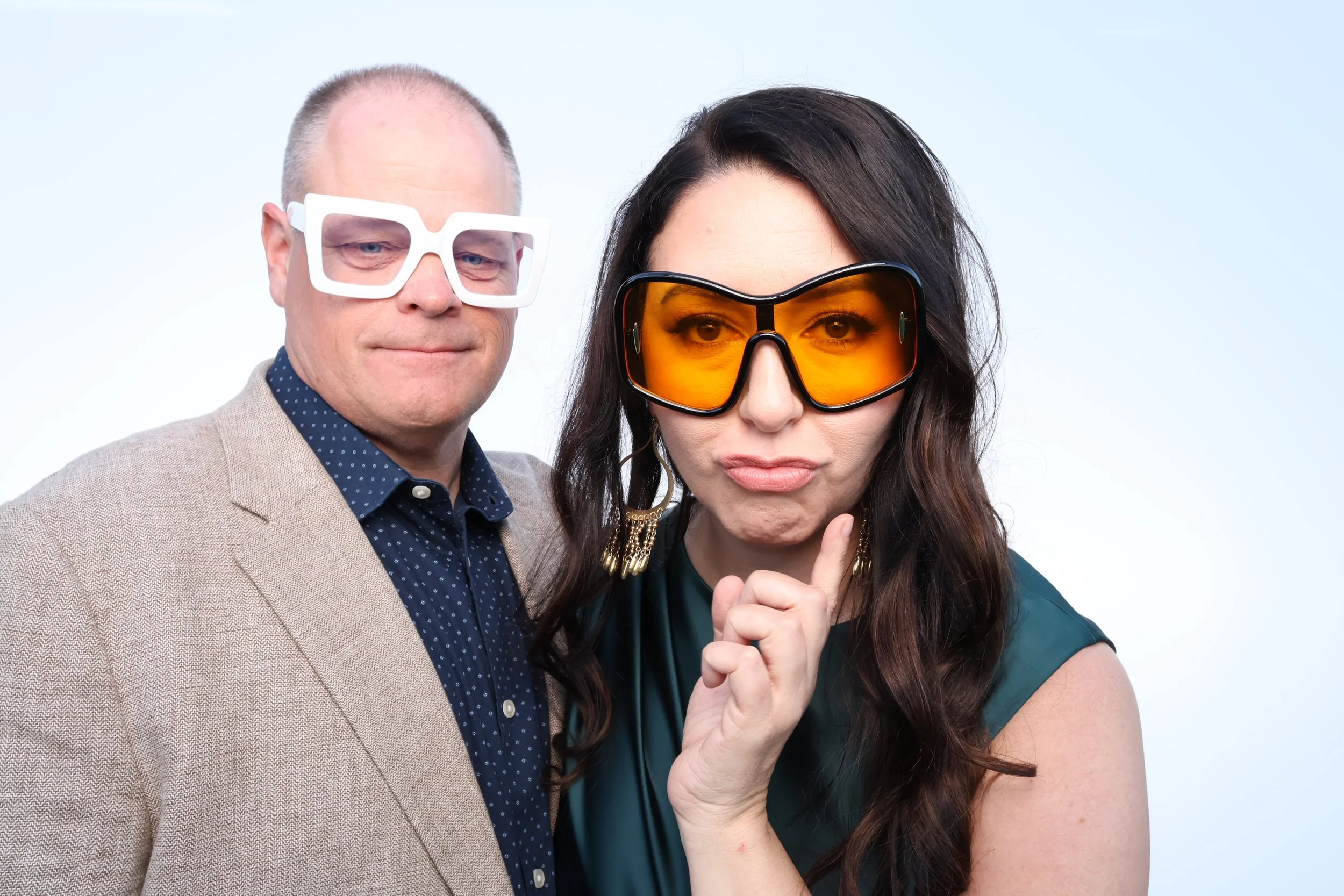 A man and a woman wearing large sunglasses, standing close together against a plain light background.