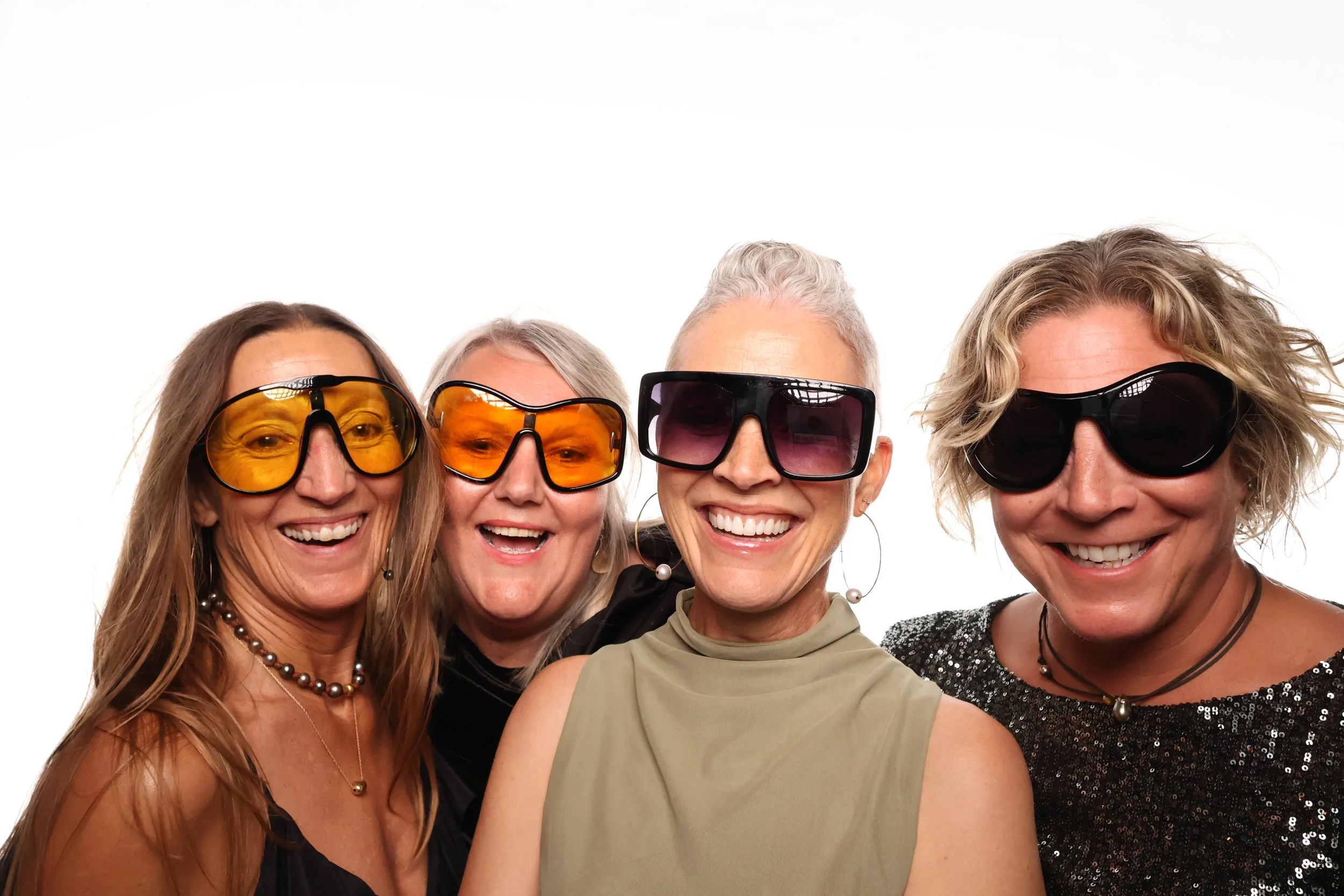 Four smiling women wearing sunglasses, posing together against a white background.