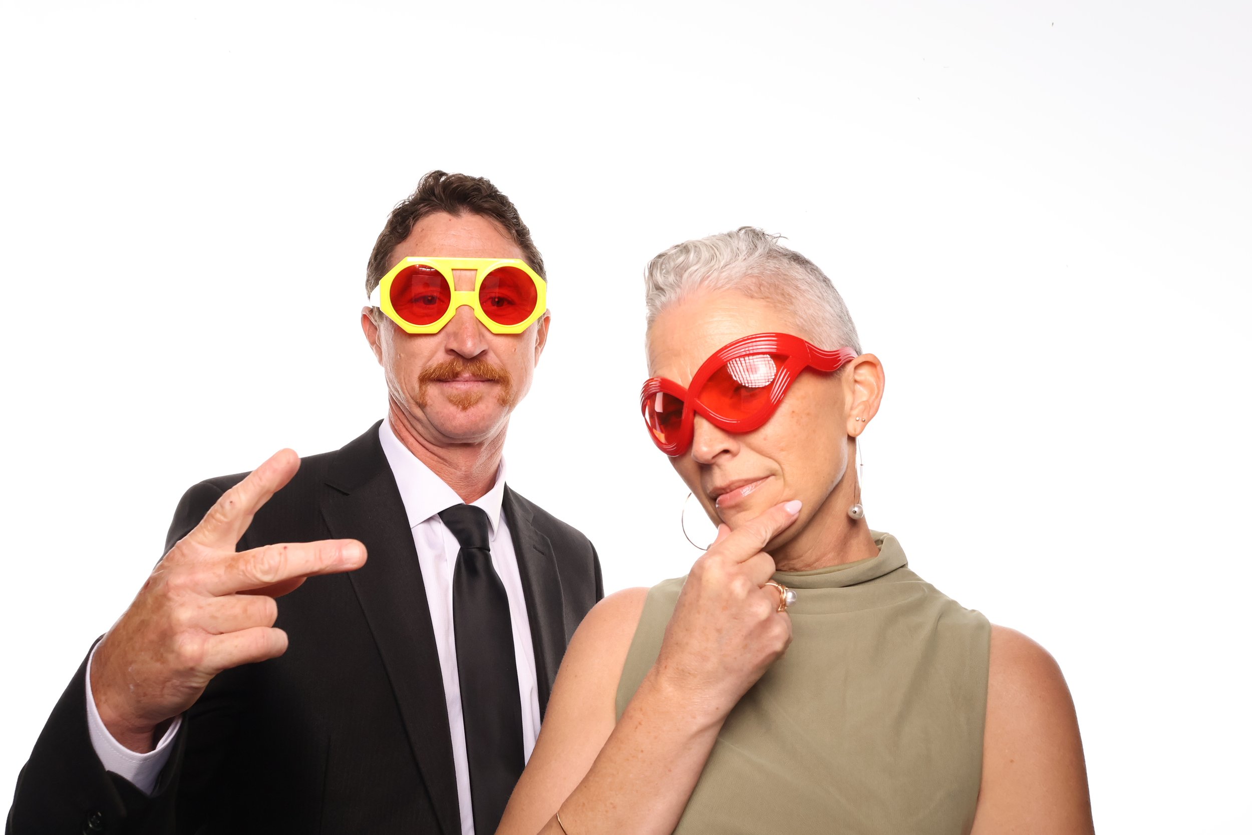A man in a black suit and tie wearing yellow sunglasses with red lenses, making a peace sign, standing next to a woman with short gray hair wearing red sunglasses, touching her chin with her hand, both against a white background.