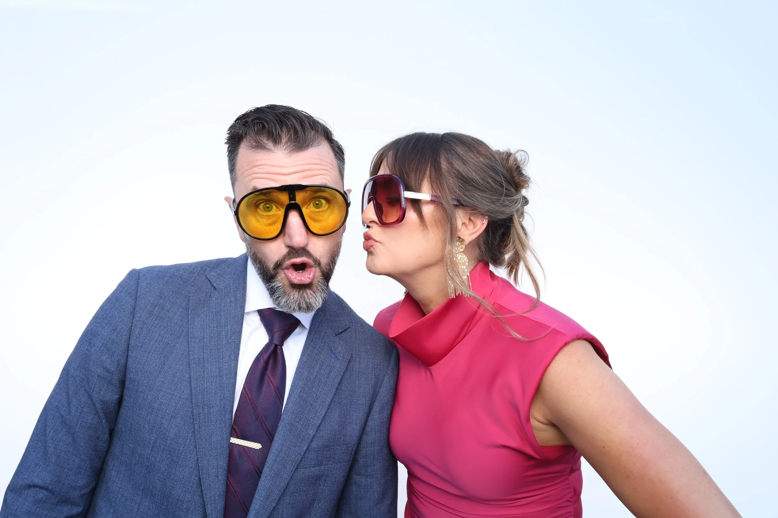 A man and woman posing together; the man is wearing a blue suit with a white shirt and patterned tie, and yellow tinted glasses, looking surprised; the woman is wearing a bright pink dress with large earrings and dark tinted glasses, leaning in to ki