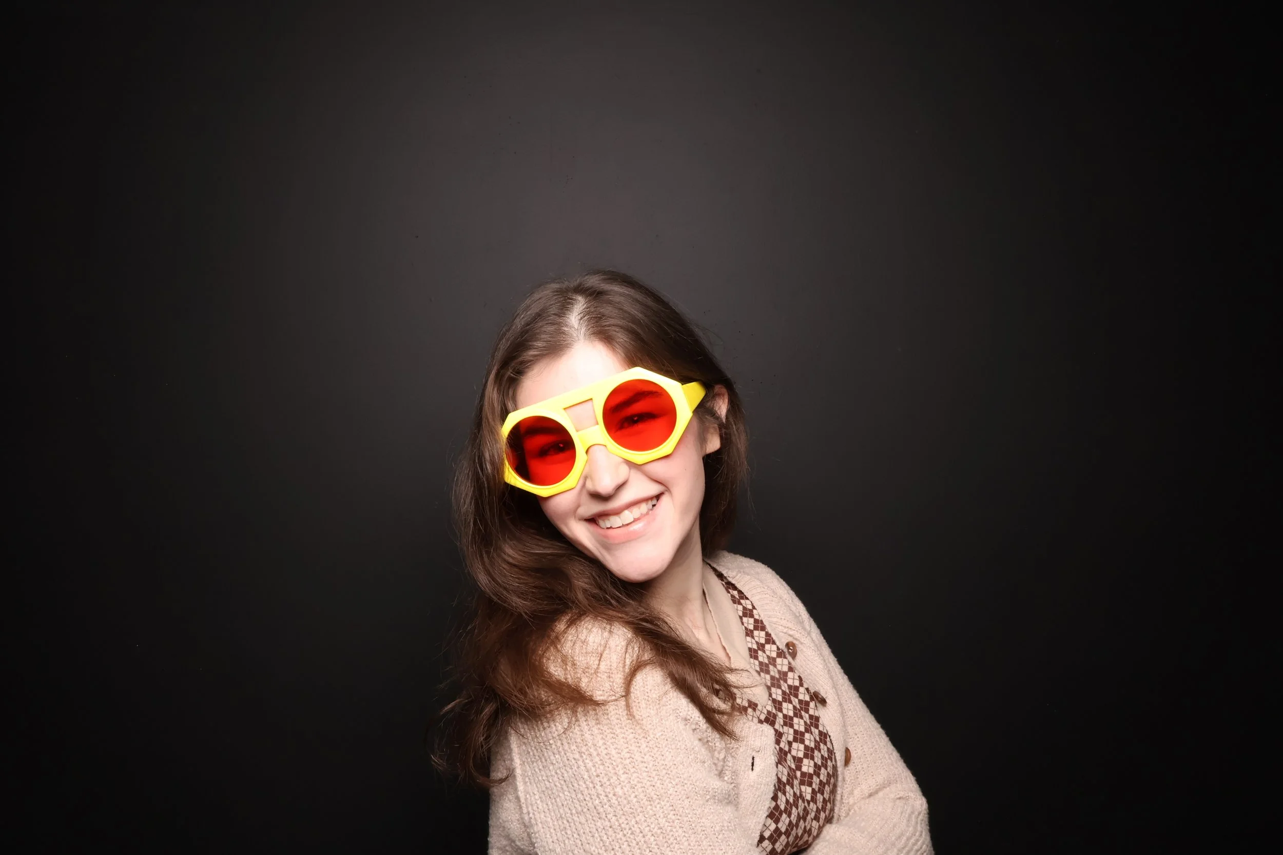 Young woman with long brown hair smiling, wearing yellow and red sunglasses, beige sweater, and patterned shirt against a black background.