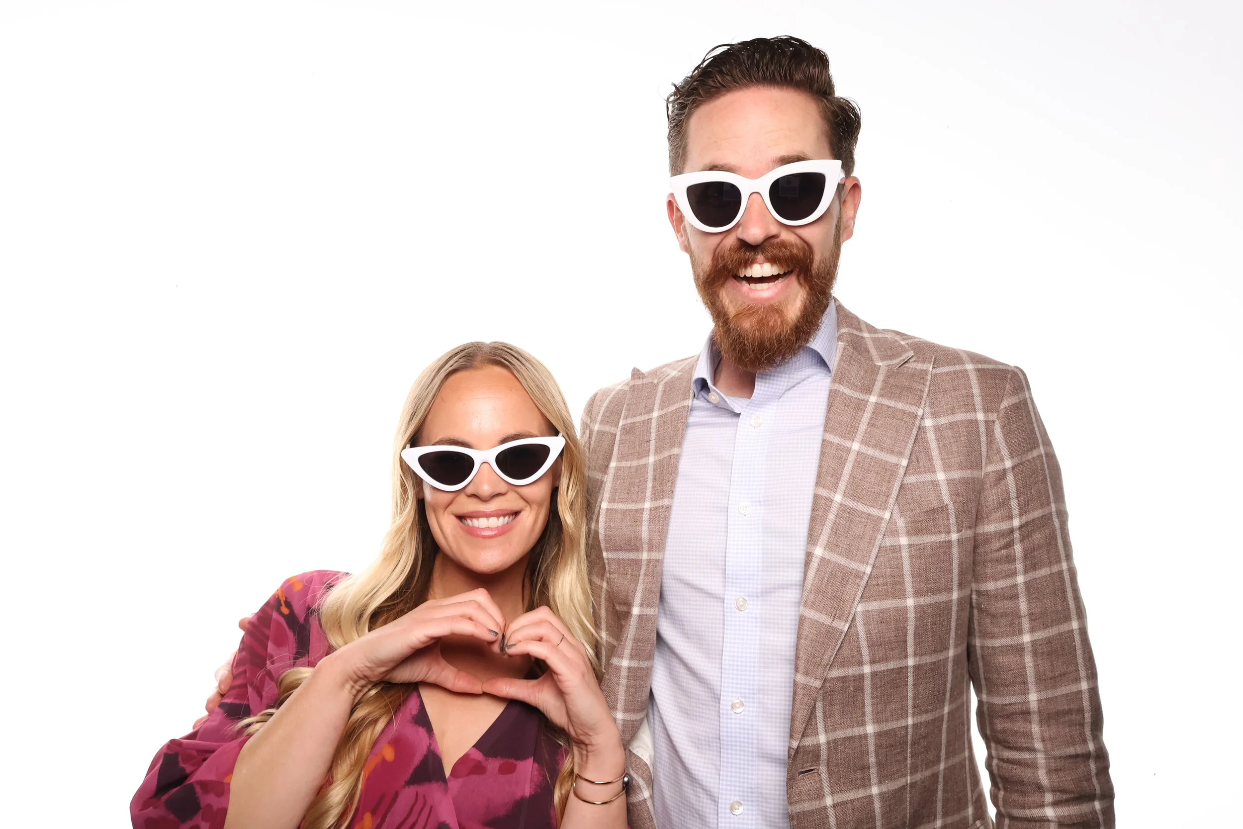 Couple photo booth pictures of a smiling woman with blonde hair and sunglasses making a heart shape with her hands, standing next to a man with a beard and sunglasses, both dressed in stylish clothing against a plain white background.