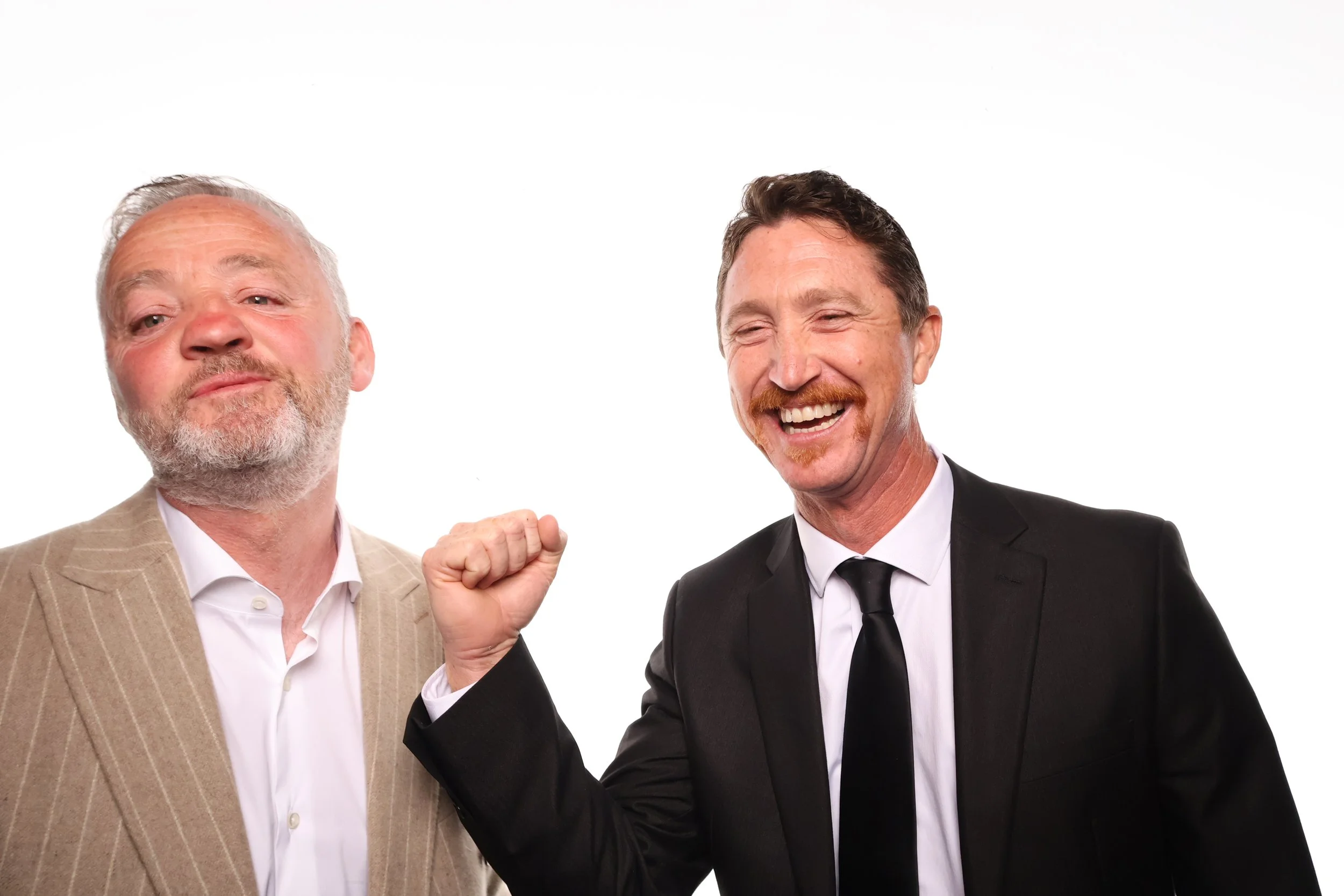 Two men in suits, one older with a beard and gray hair, raising their fists in a gesture of victory or celebration, against a plain white background.