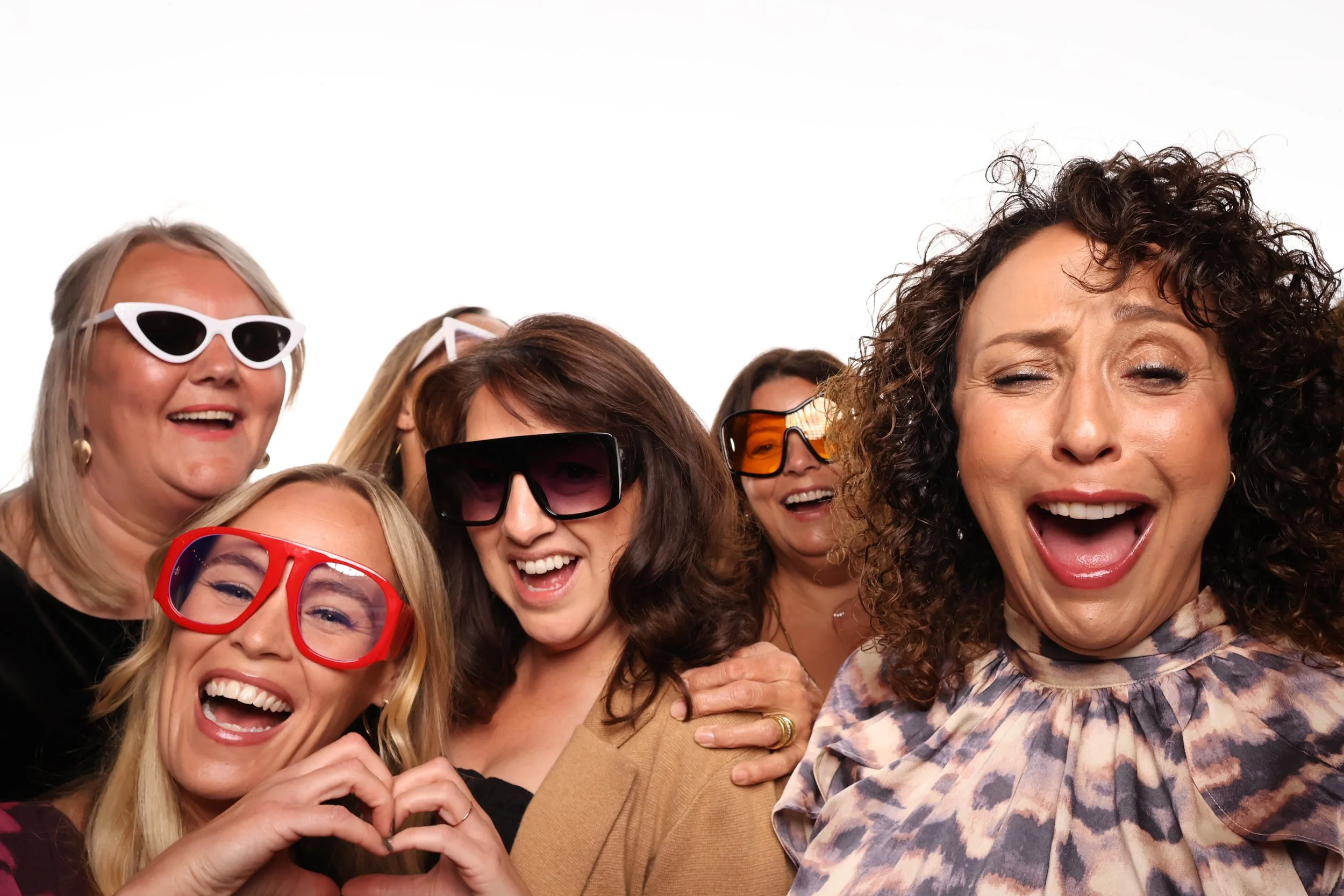 A group of women wearing colorful and stylish sunglasses, smiling and laughing together against a white background.