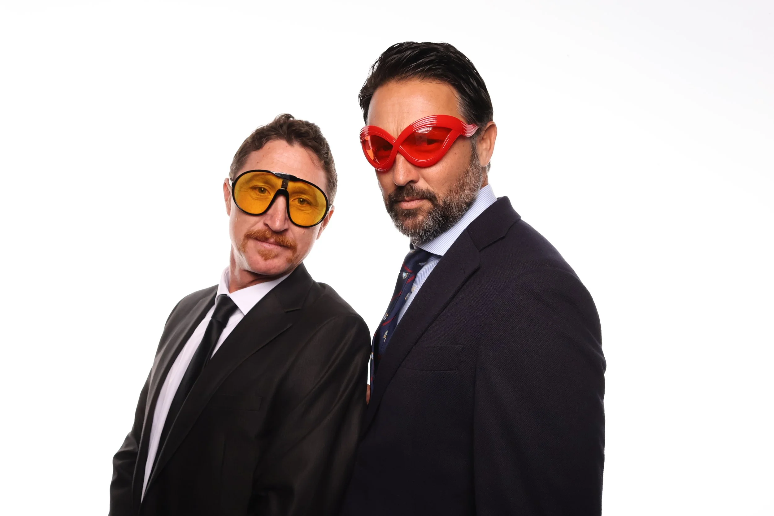 High quality booth pictures of two men in suits wearing colorful goggles, one with red glasses and the other with yellow glasses, posing against a plain white background.