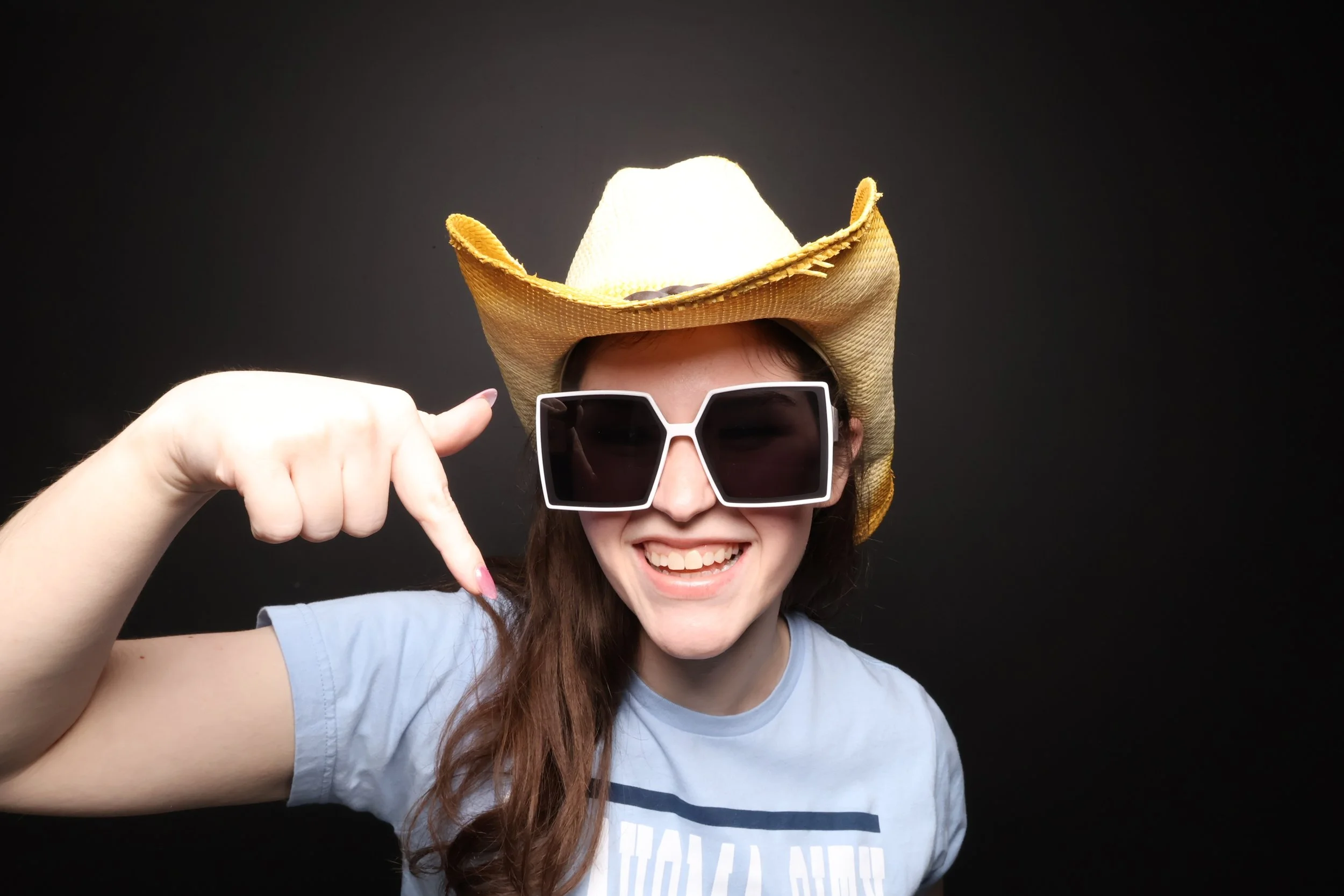 A smiling woman wearing large sunglasses and a cowboy hat pointing at her sunglasses against a black background.