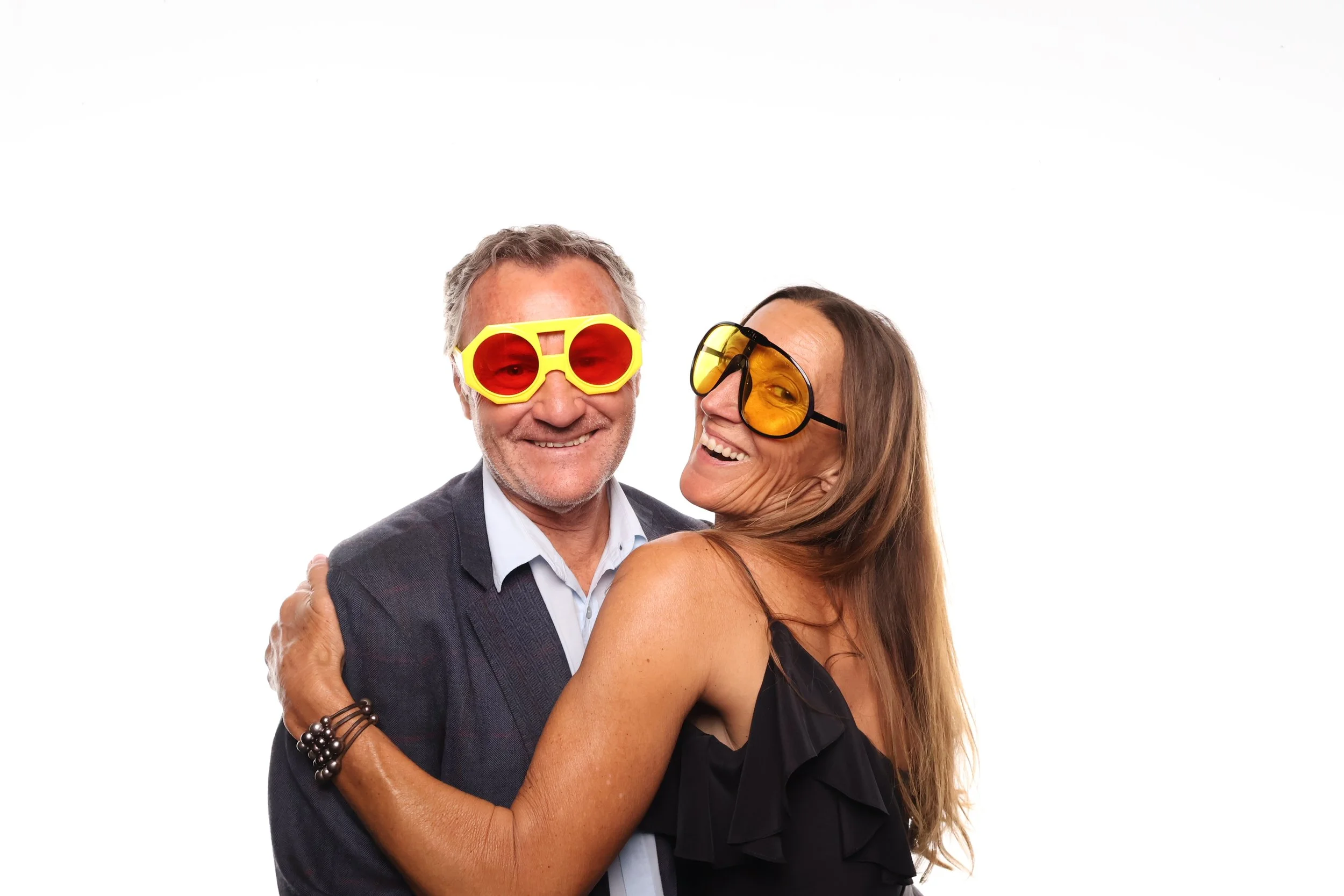 A man with gray hair and a beard wearing yellow and red sunglasses, smiling, embracing a woman with long brown hair wearing orange-tinted glasses and a black dress, both smiling and looking happy.