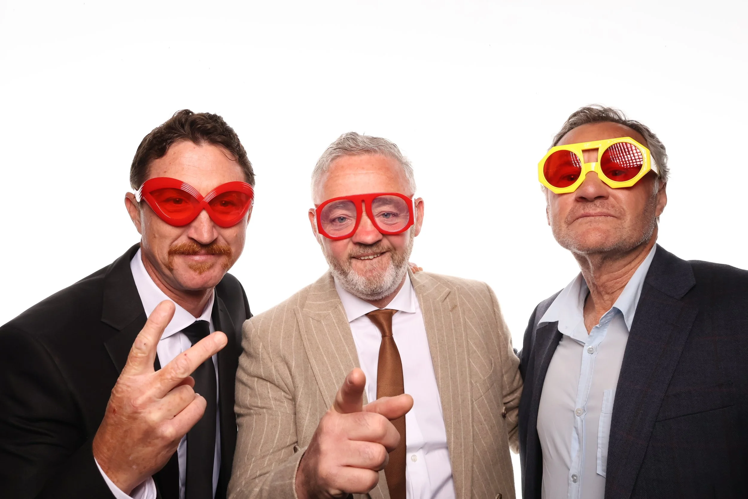 Sick booth pictures of three men dressed in suits wearing colorful sunglasses, smiling and making peace signs against a white background.