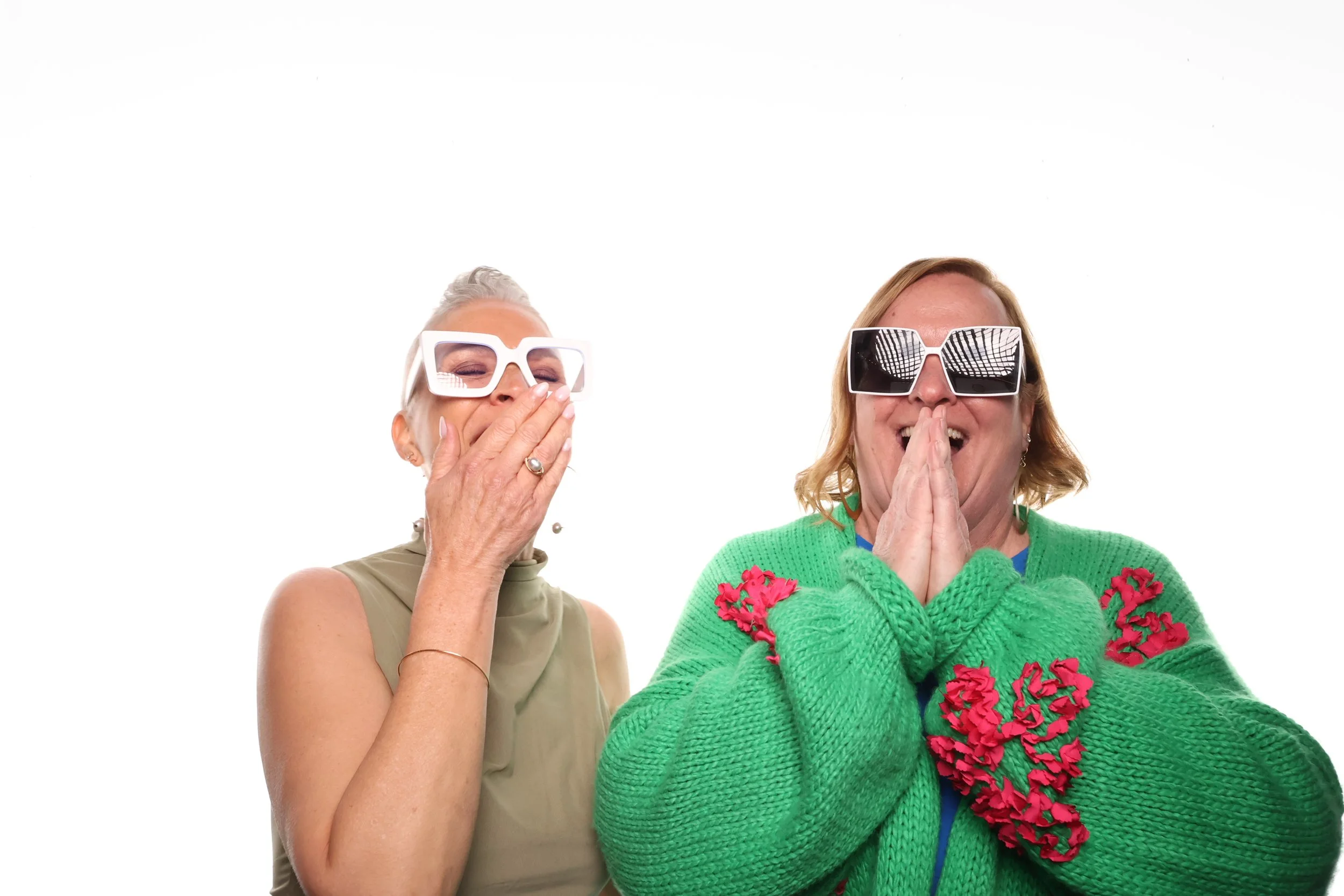 Two women are smiling and laughing, wearing large fashion sunglasses. One woman has short gray hair and is covering her mouth with her hand. The other woman has blond hair, is wearing a green sweater with red floral accents, and has her hands pressed