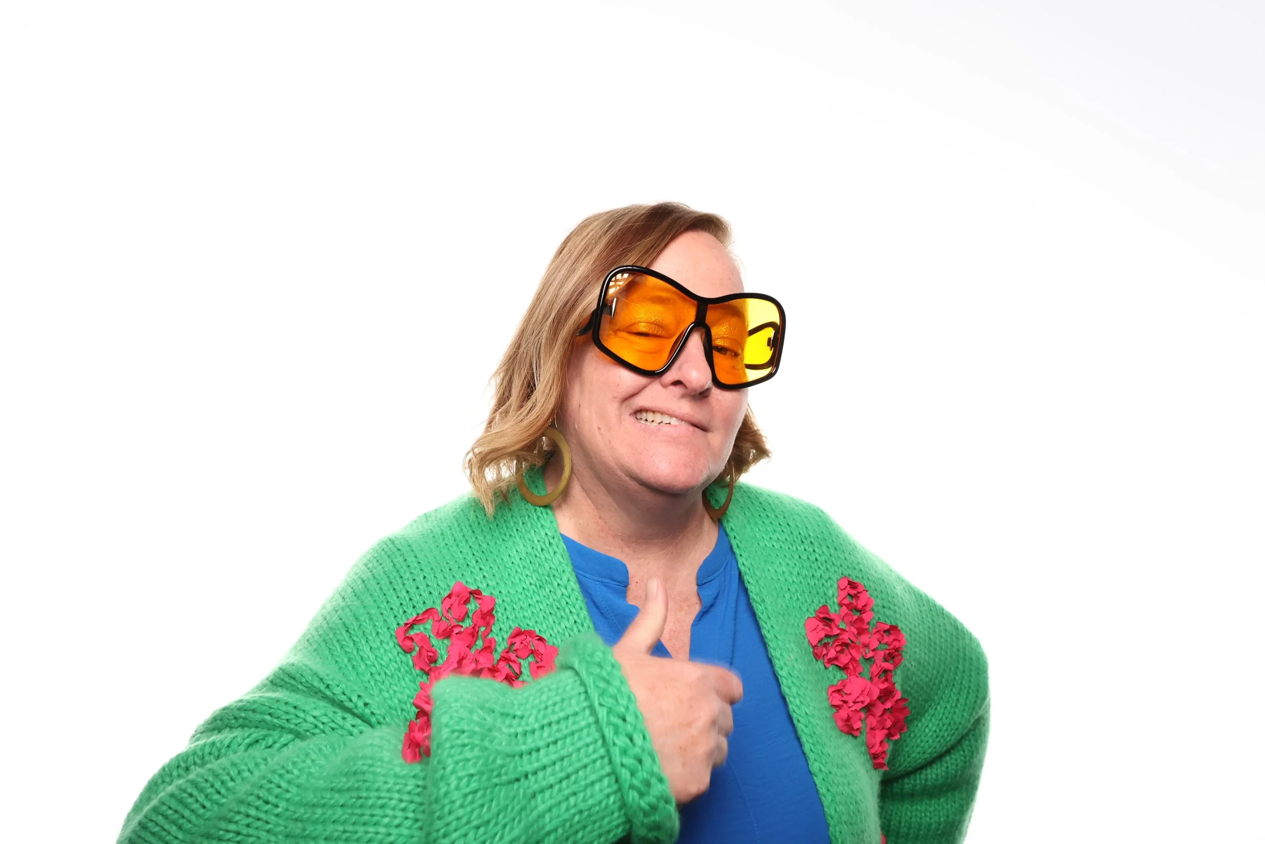 A woman wearing orange tinted glasses and a bright green cardigan with pink floral accents, giving a thumbs-up gesture.