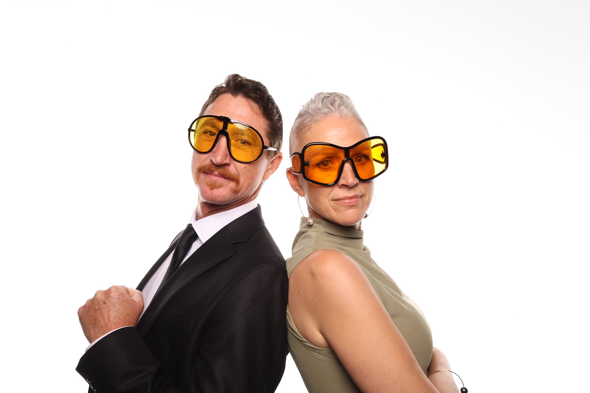 A man in a suit with yellow-tinted sunglasses and a woman in a beige top with yellow-tinted sunglasses, stand back-to-back against a white background.