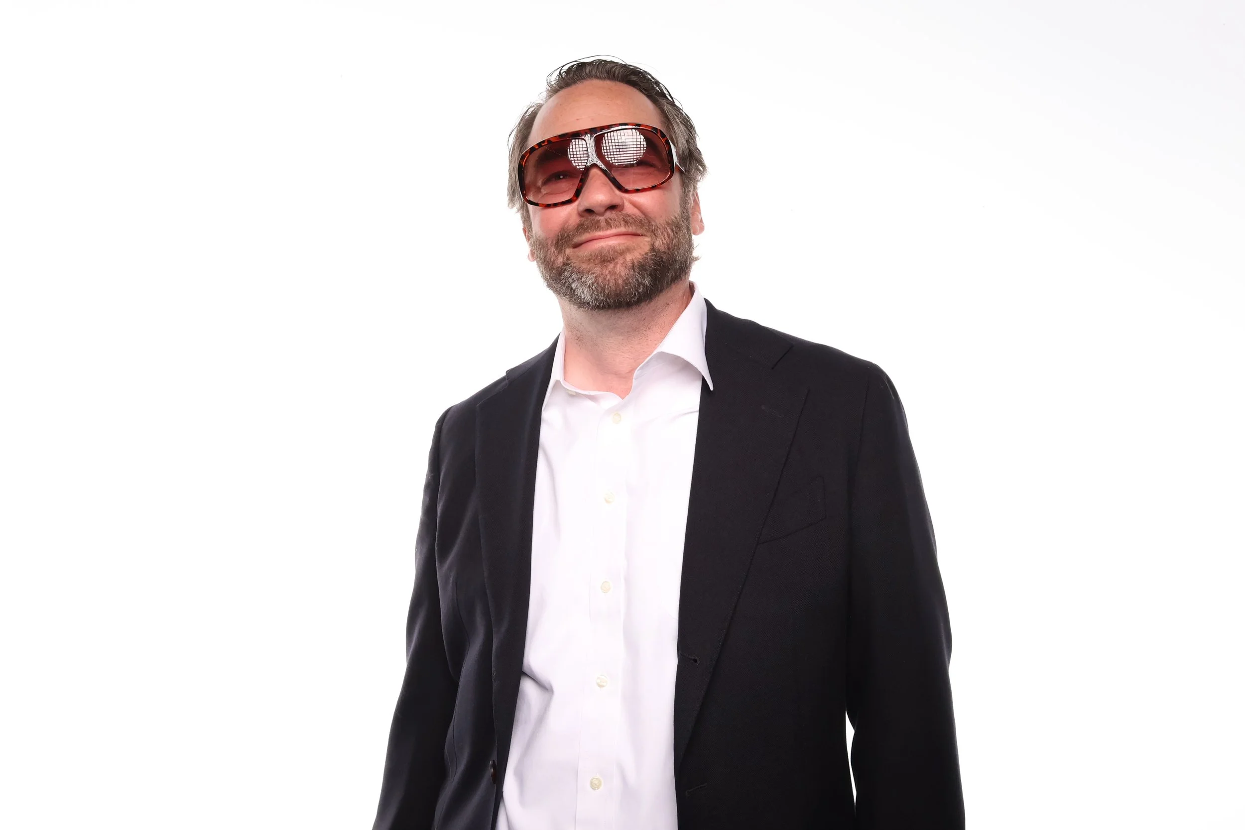 Man with gray hair and beard wearing a black suit, white shirt, and sunglasses, standing against a white background.