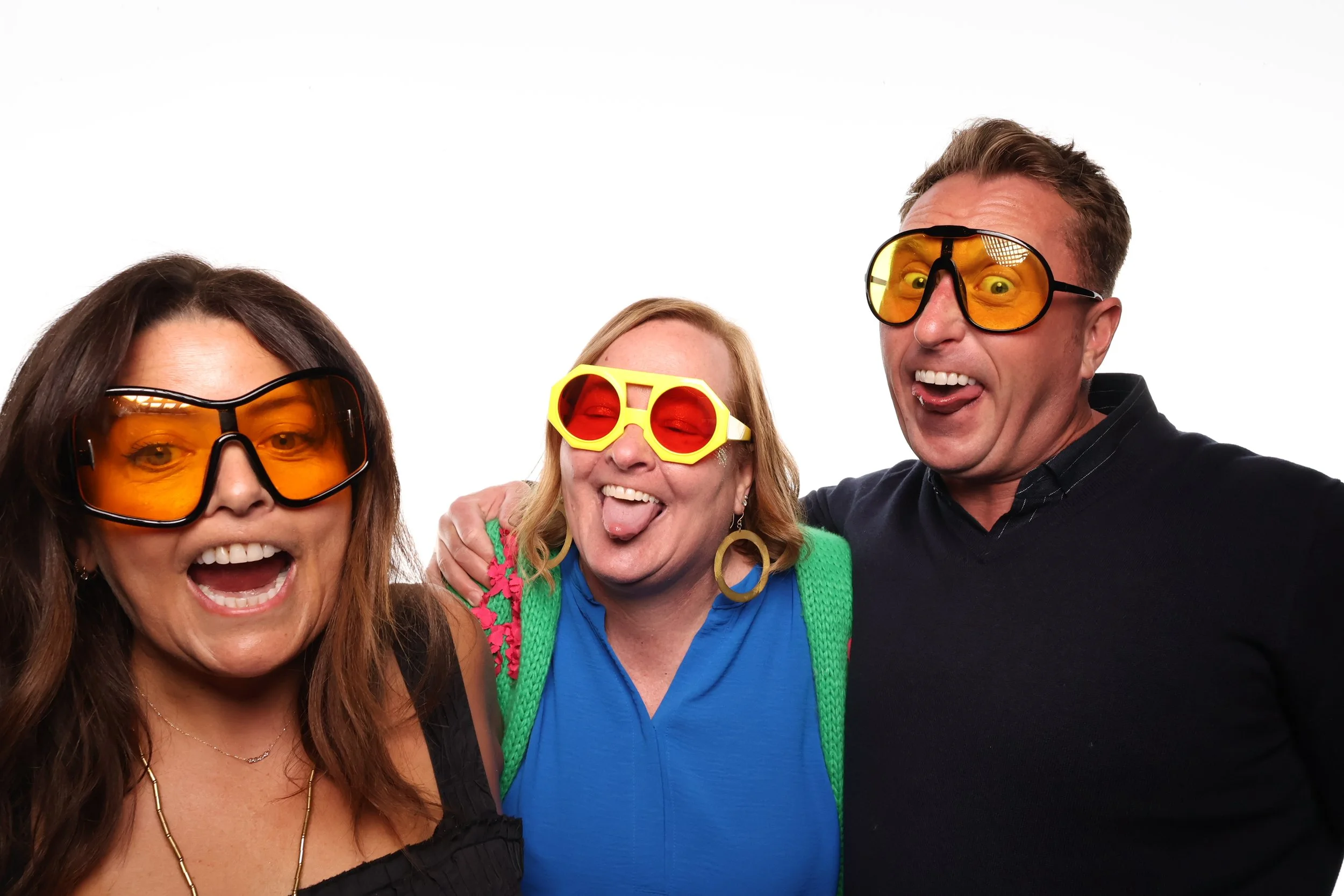 Three people having fun, wearing colorful sunglasses, smiling and making silly faces, against a white background.